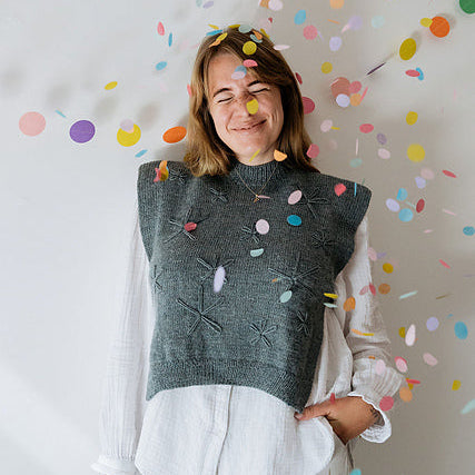 Woman wearing a dark gray sleeveless sweater with colorful patterns against a white background with confetti.