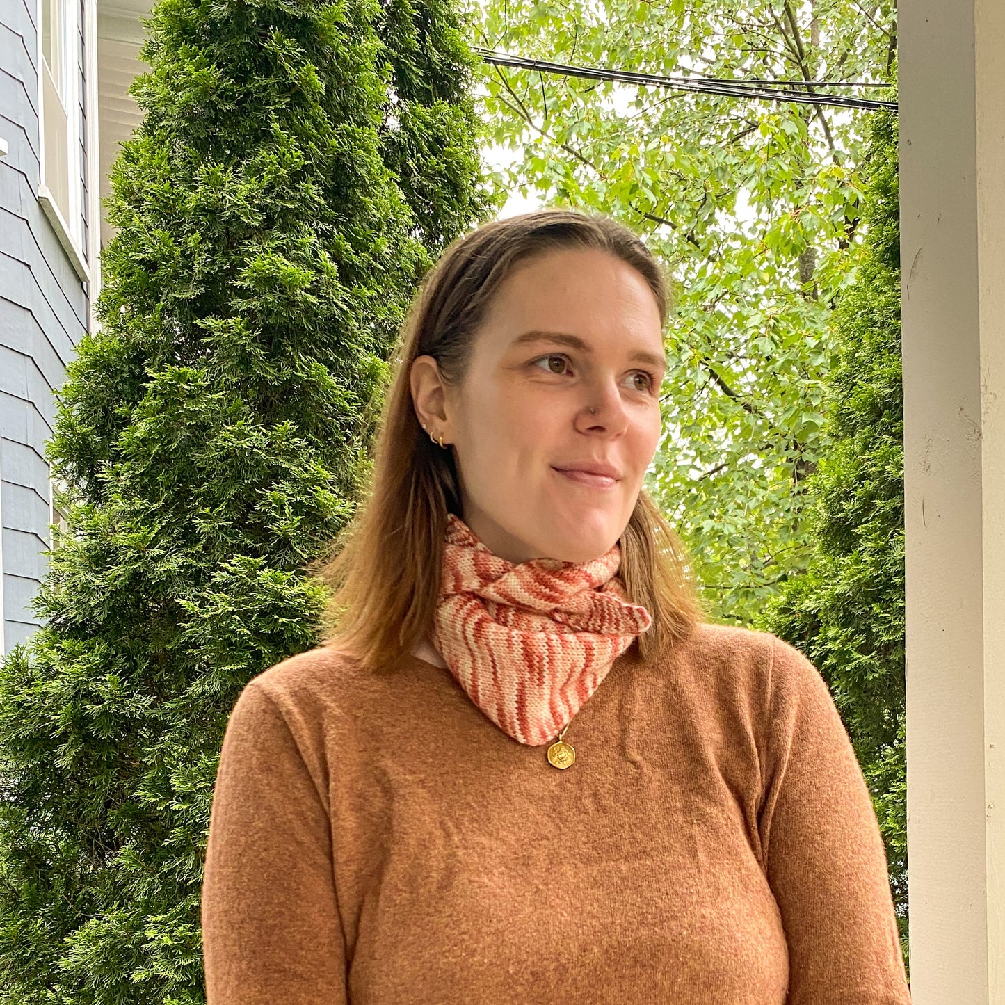 Woman wearing a brown sweater and variegated pink knitted scarf standing outdoors with greenery in the background