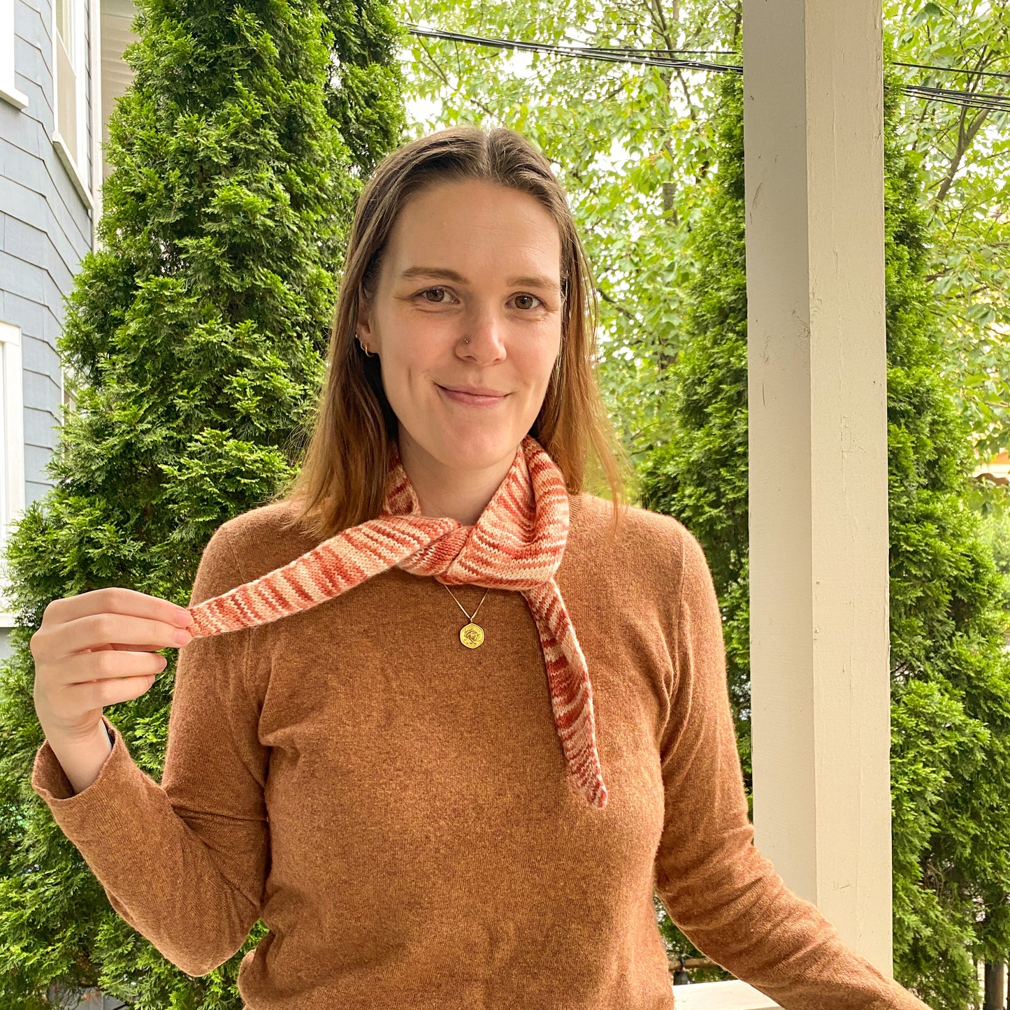 Woman wearing a brown sweater and variegated knitted bandana scarf on a porch with greenery in the background