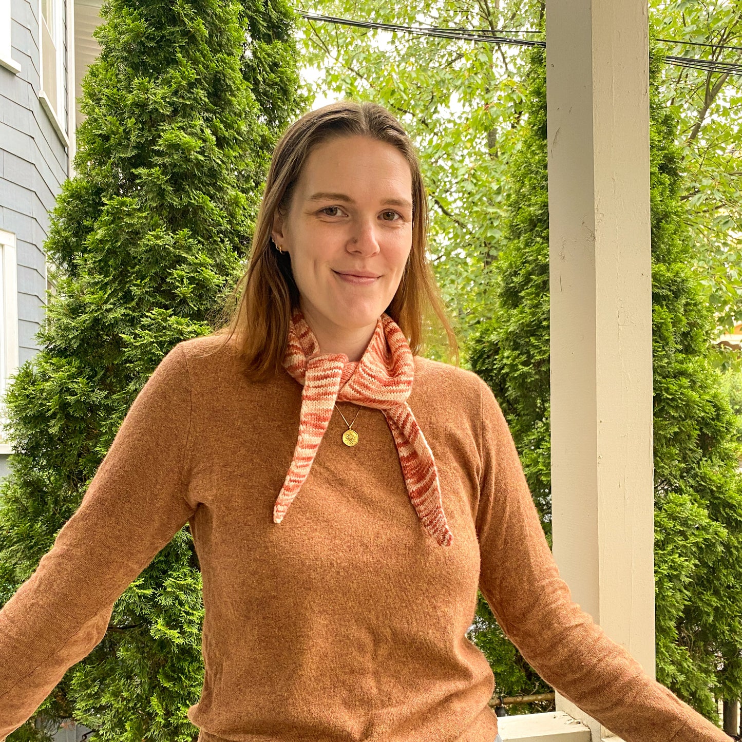Woman standing on a porch wearing a brown sweater and variegated knitted bandana scarf, with greenery in the background.