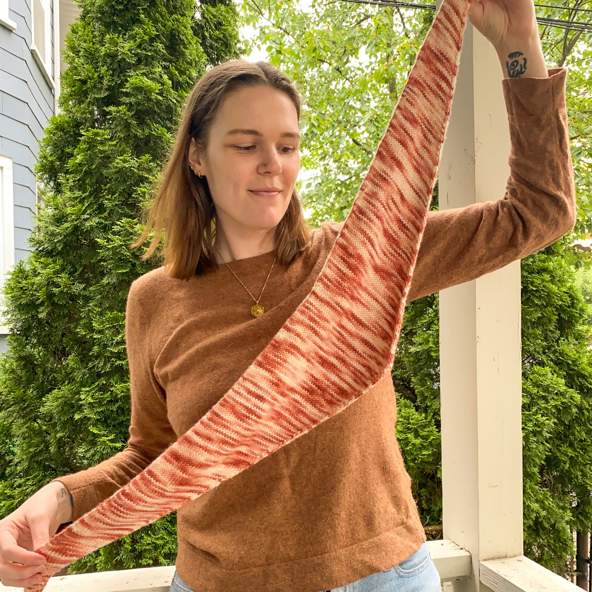 Person holding a variegated pink knitted triangle scarf on a porch with greenery in the background