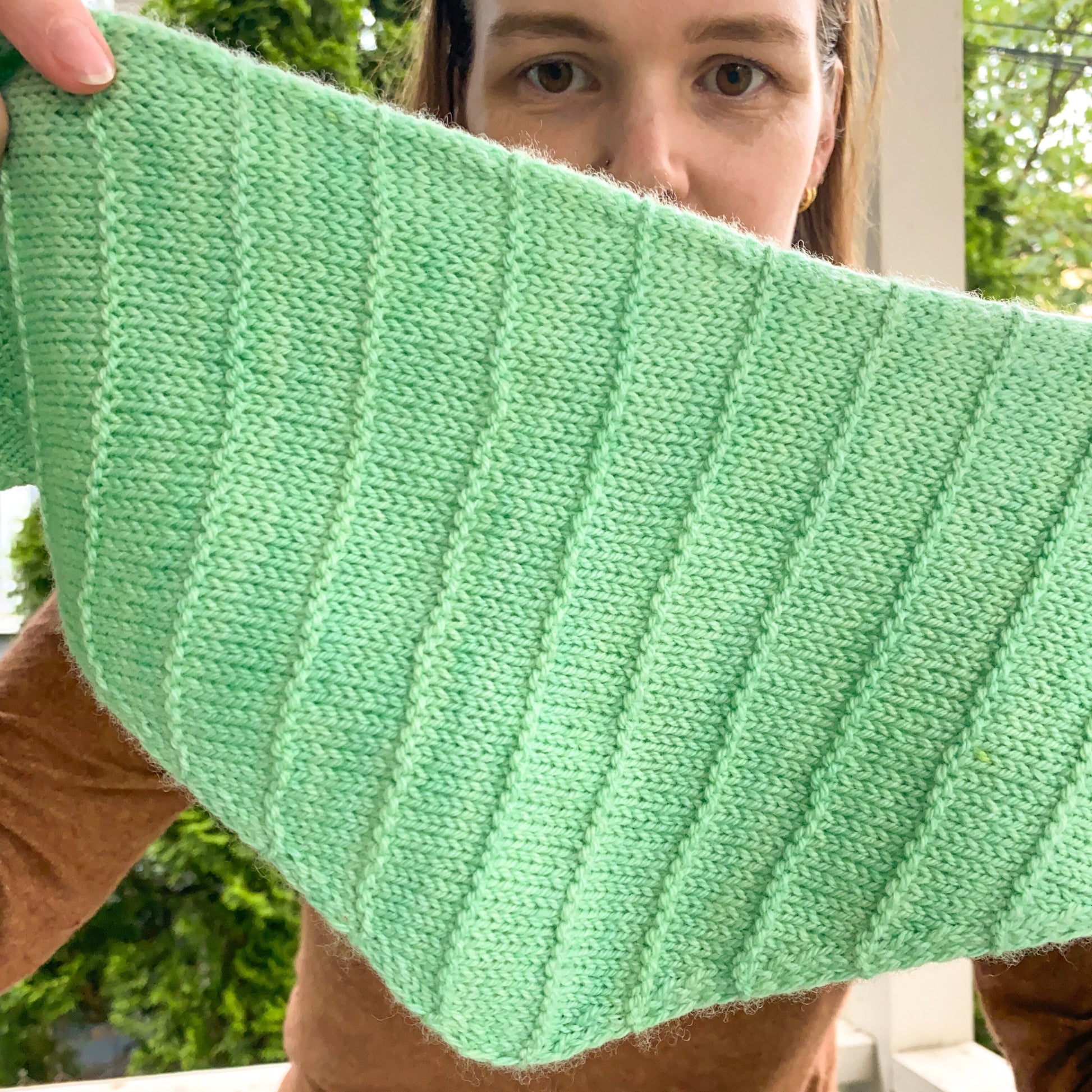 Person holding a green knitted triangle scarf with purl ridge stripes outdoors