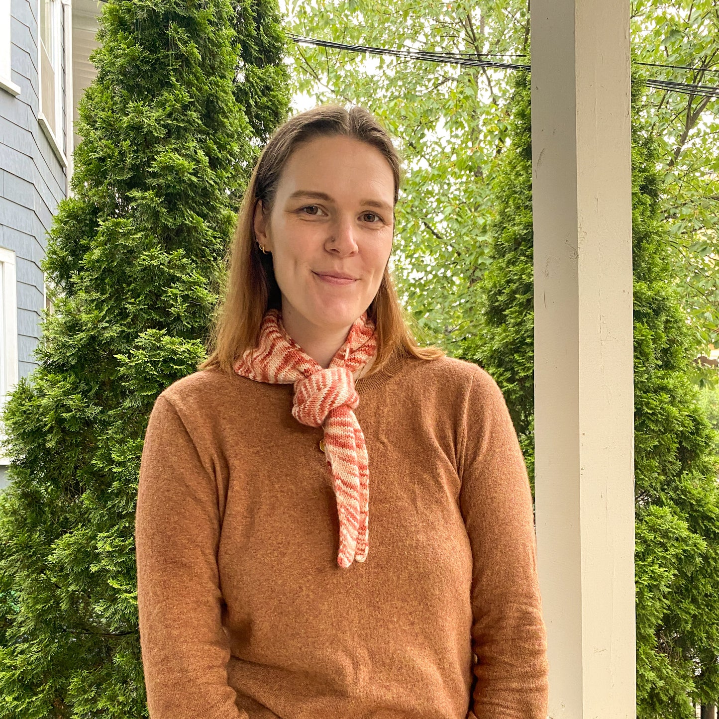 Woman wearing a brown sweater and variegated pink bandana scarf standing on a porch with greenery in the background