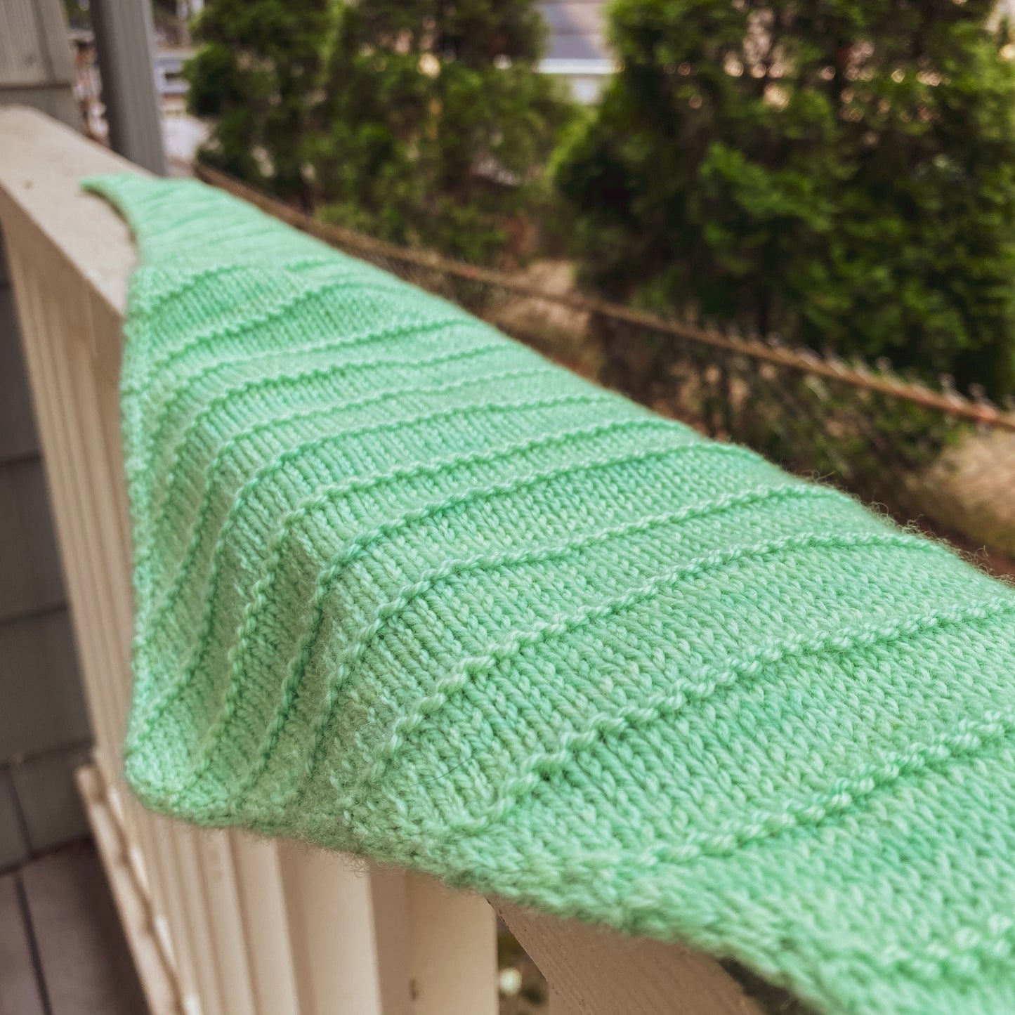 Green knitted triangle scarf with purl ridge stripes draped over a railing with trees in the background
