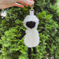 Hand holding a sheep-shaped keychain against a green tree background