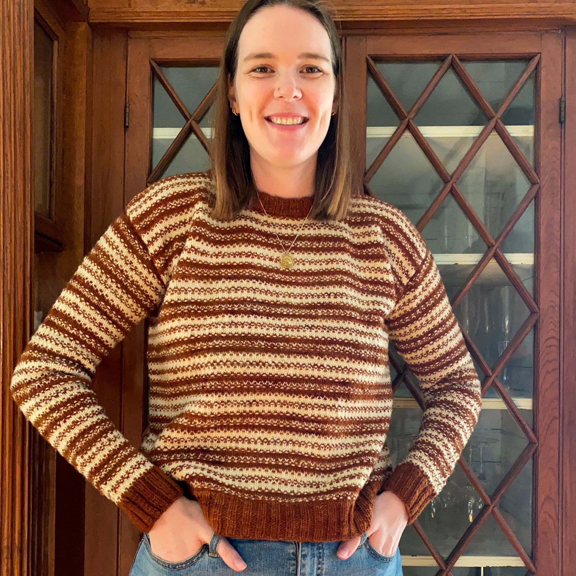 Woman wearing a rust orange and cream mosaic knitted sweater standing in front of a wooden cabinet.