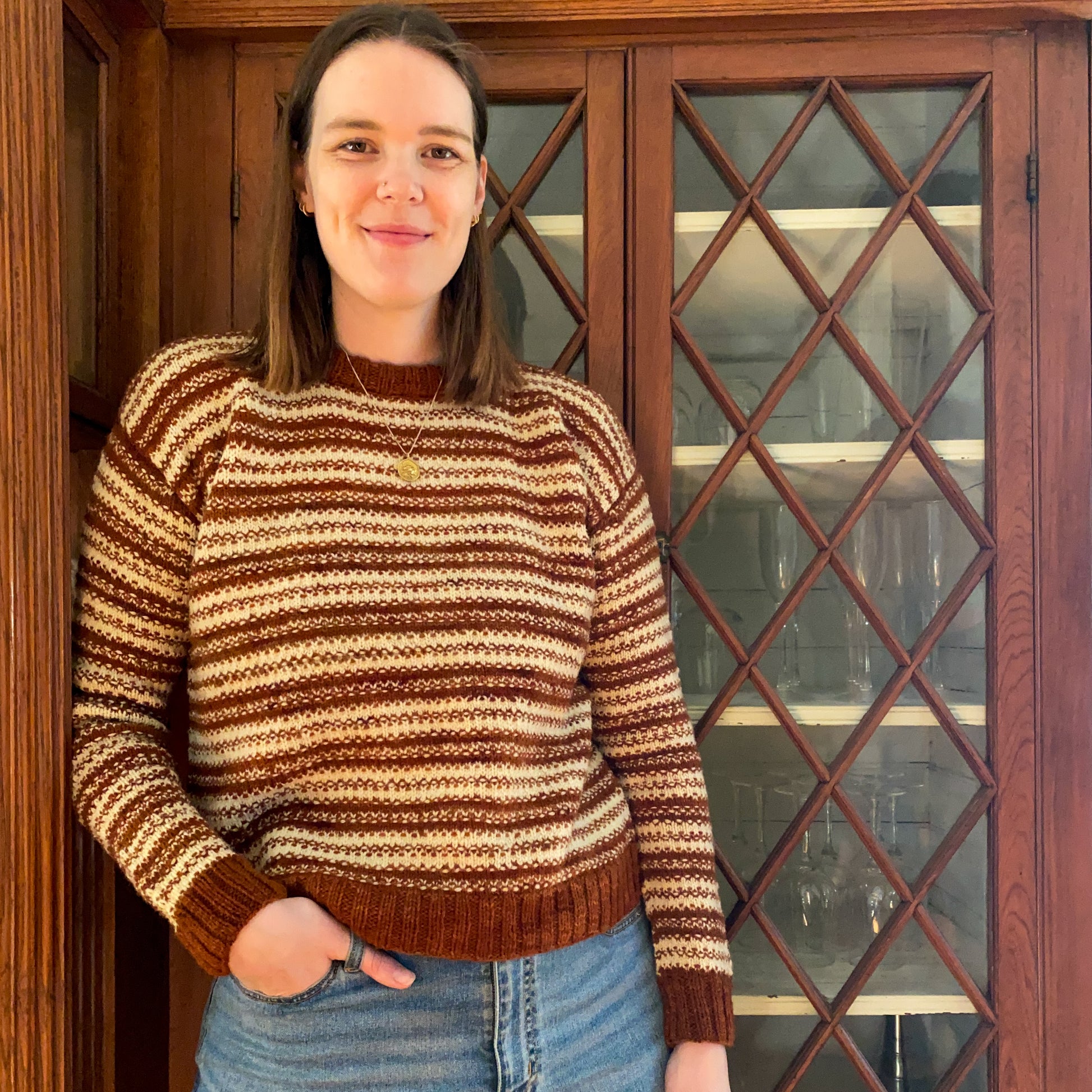 Woman wearing a rust orange and cream mosaic knitted sweater standing in front of a wooden cabinet.