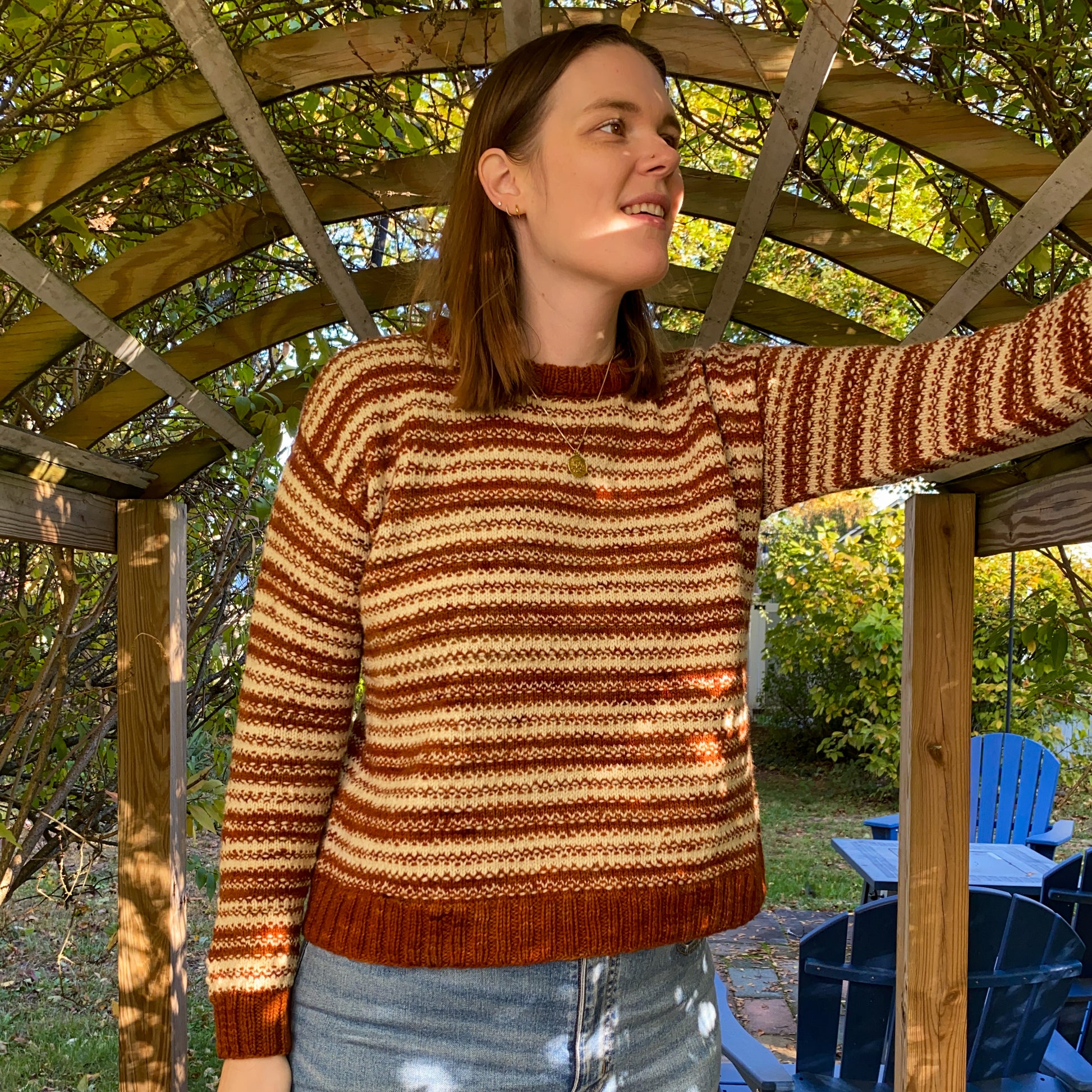 Woman wearing a striped knitted sweater standing under a wooden arbor with greenery in the background