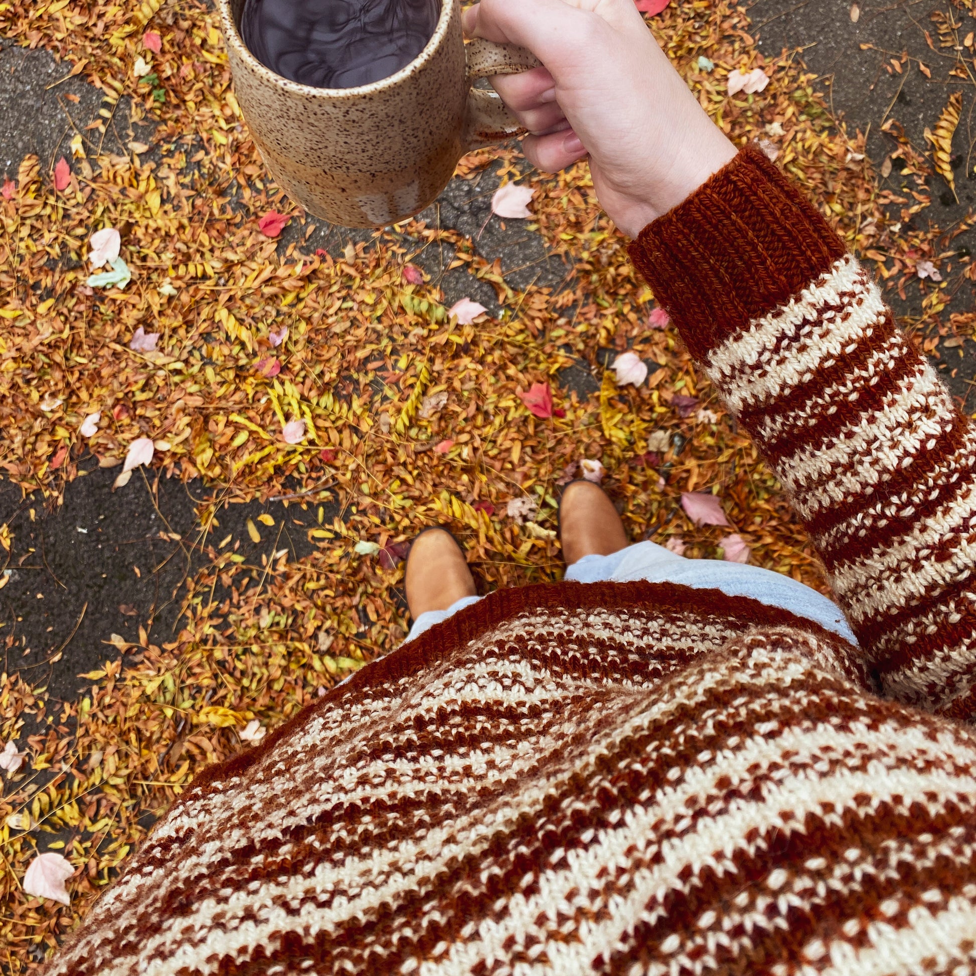 Person wearing a rust orange and cream knitted sweater and holding a mug of coffee on a ground covered with autumn leaves