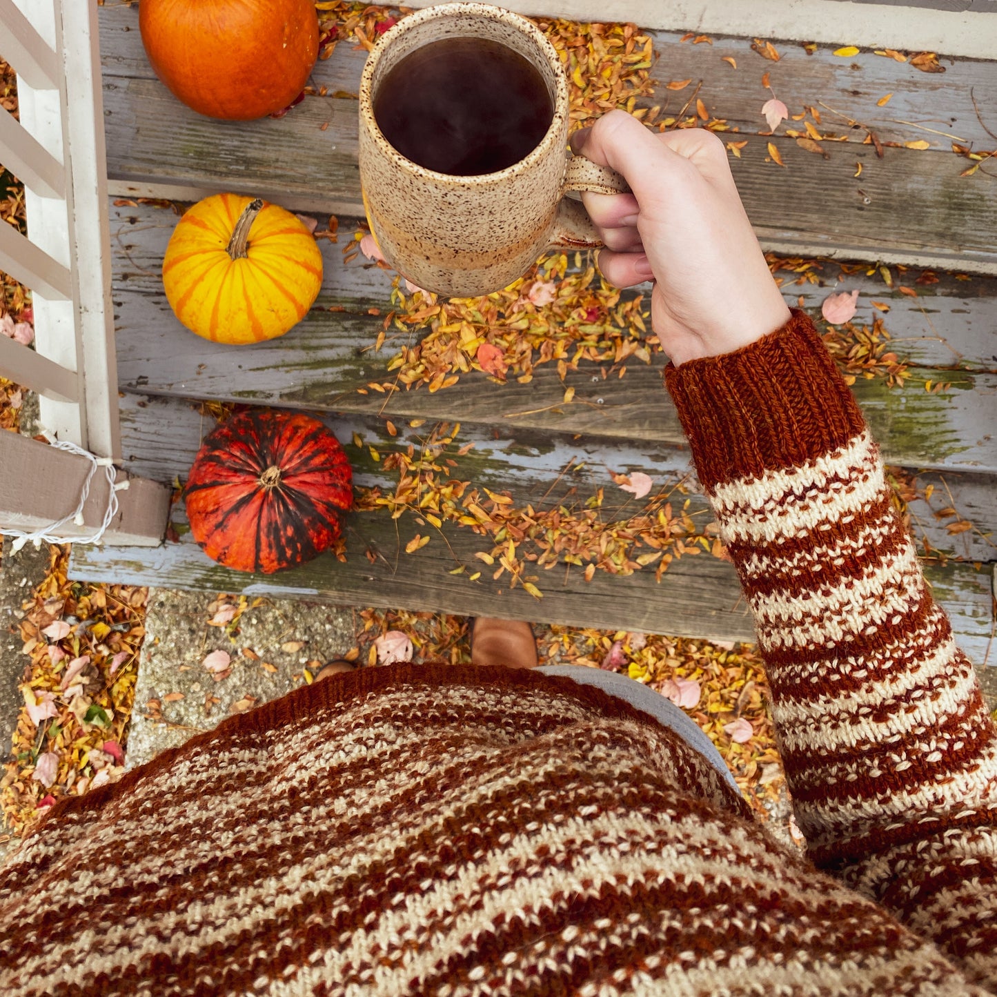 Person wearing a rust orange and cream knitted sweater and holding a mug of coffee on a wooden deck with pumpkins and autumn leaves.