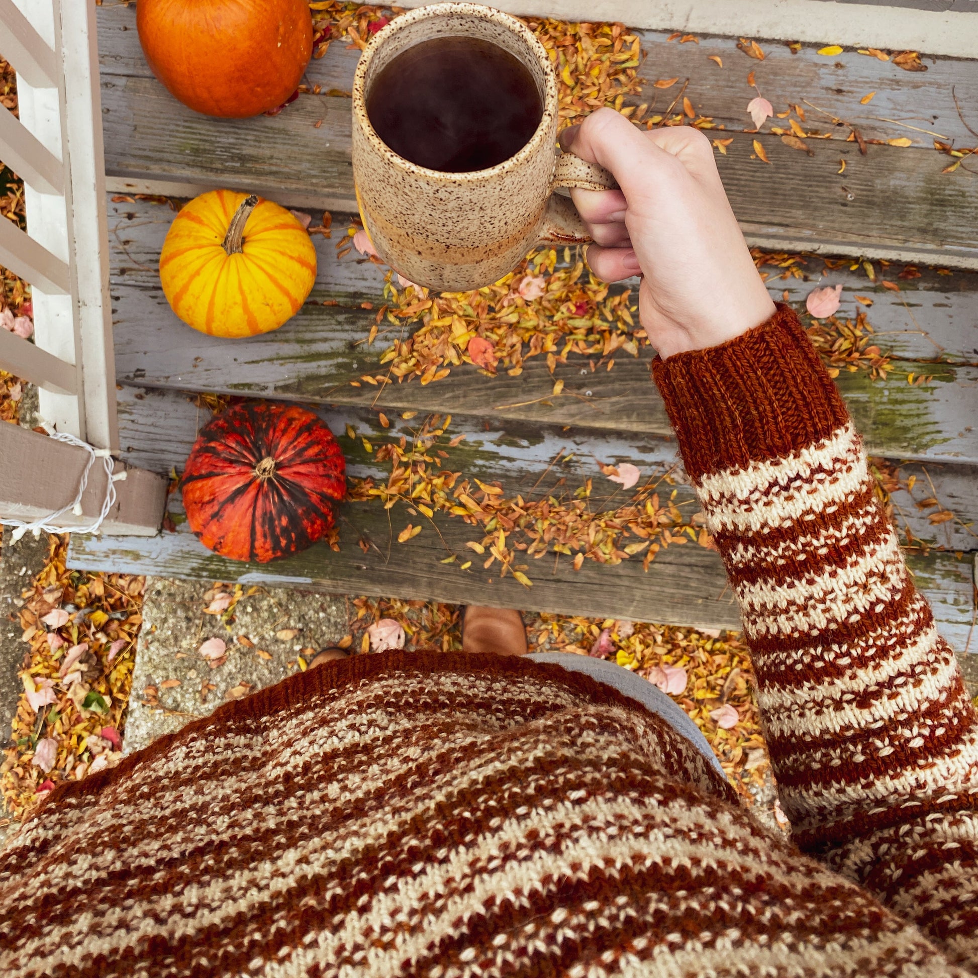 Person wearing a rust orange and cream knitted sweater and holding a mug of coffee on a wooden deck with pumpkins and autumn leaves.