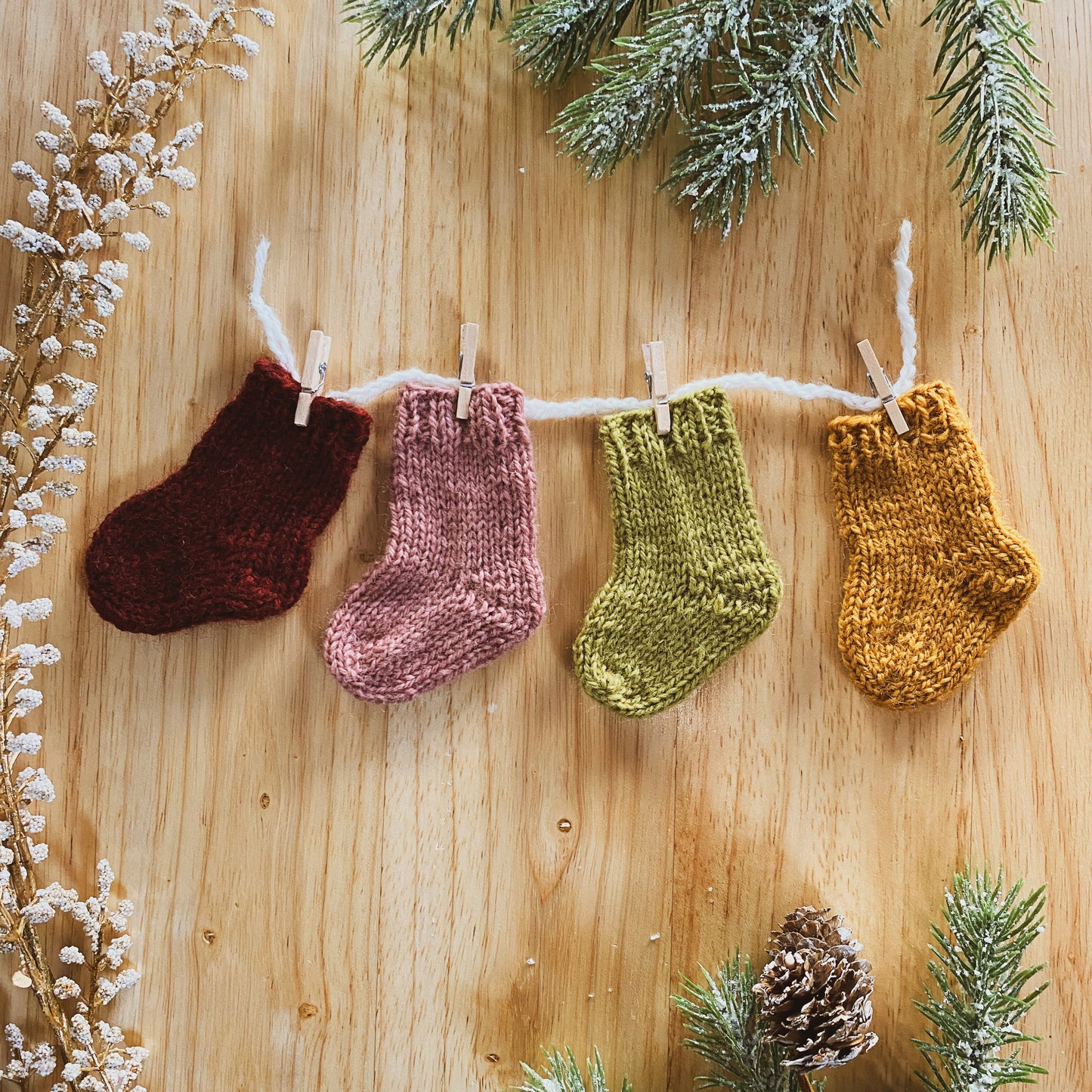 Four knitted mini socks in maroon, pink, green, and yellow hanging on a line with Christmas decorations on a wooden background.