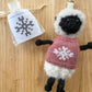 Knitted sheep keychain with pink sweater featuring a snowflake design on a wooden surface.