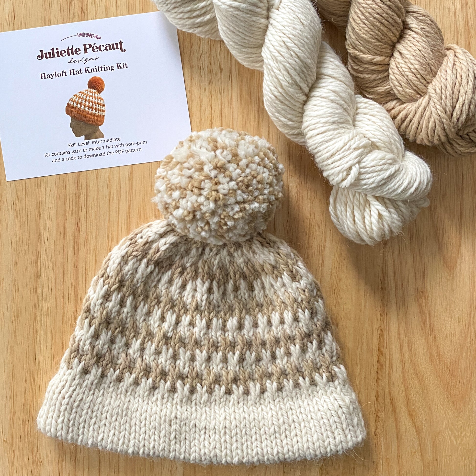 Knitted hat with pom-pom and yarn on a wooden surface, featuring Juliette Pécault Designs branding.