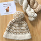 Knitted hat with pom-pom and yarn on a wooden surface, featuring Juliette Pécault Designs branding.