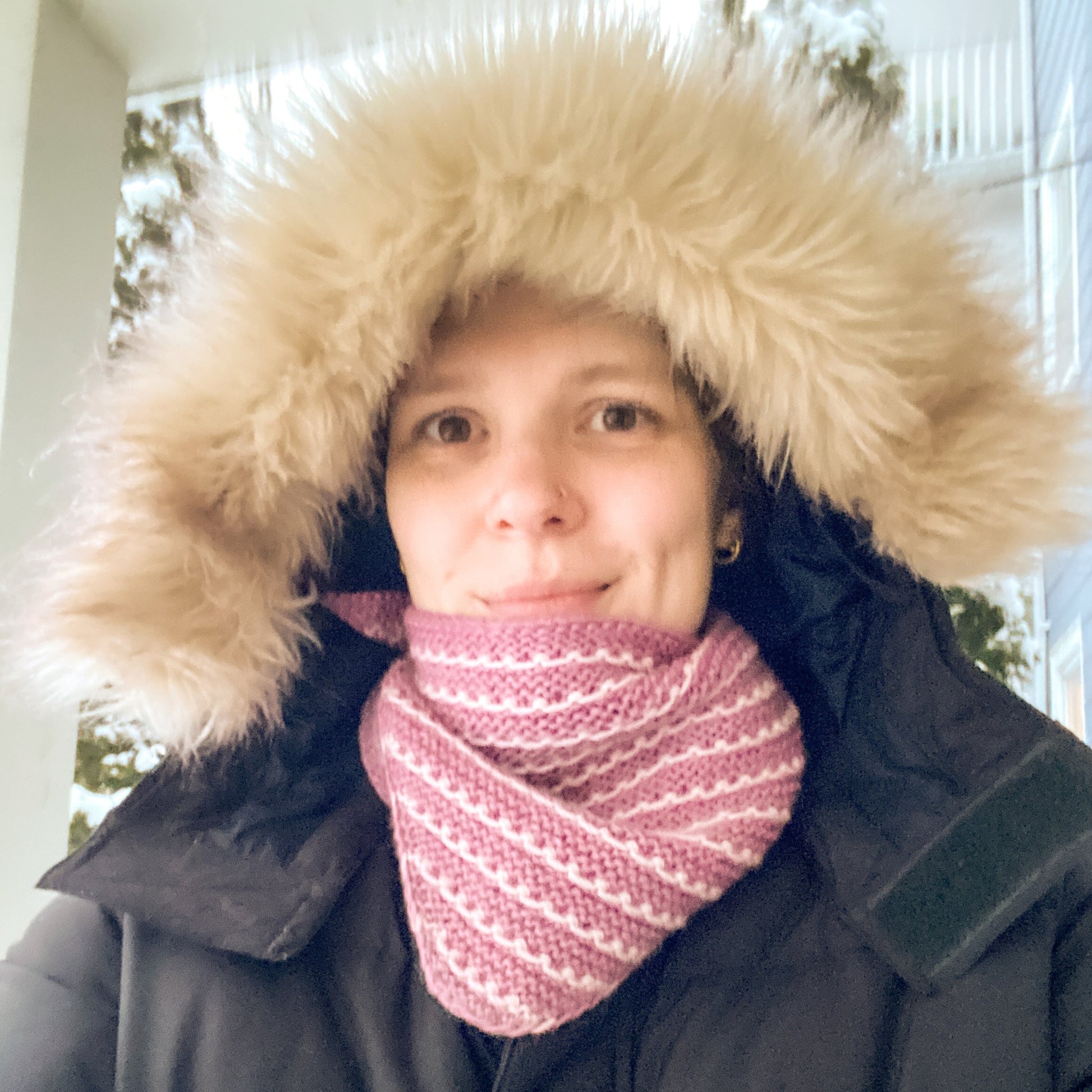 Person wearing a black coat with a fur-lined hood and a pink and white striped bandana, standing outside in the snow.