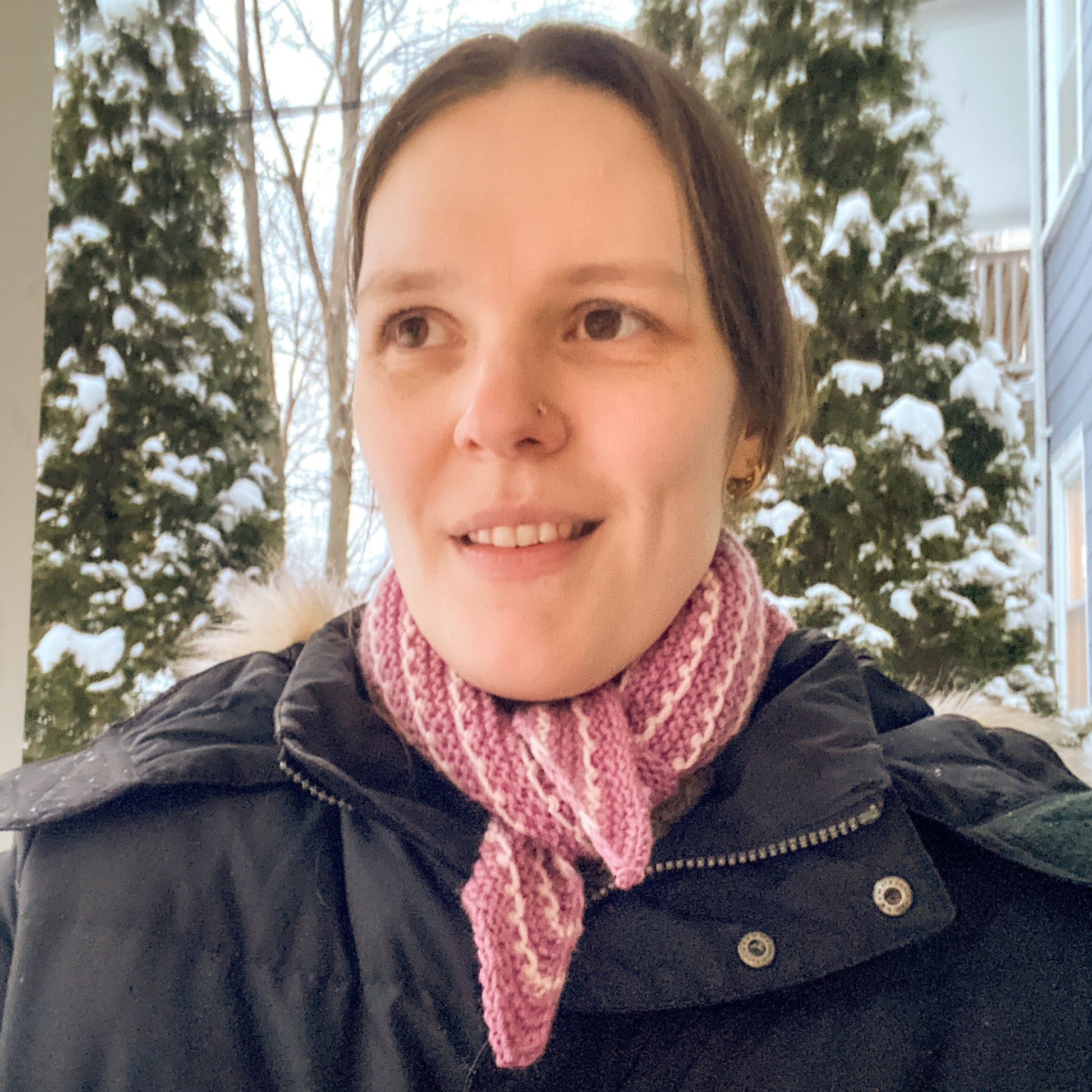 Person wearing a black coat and pink and white striped bandana outdoors with snow-covered trees in the background