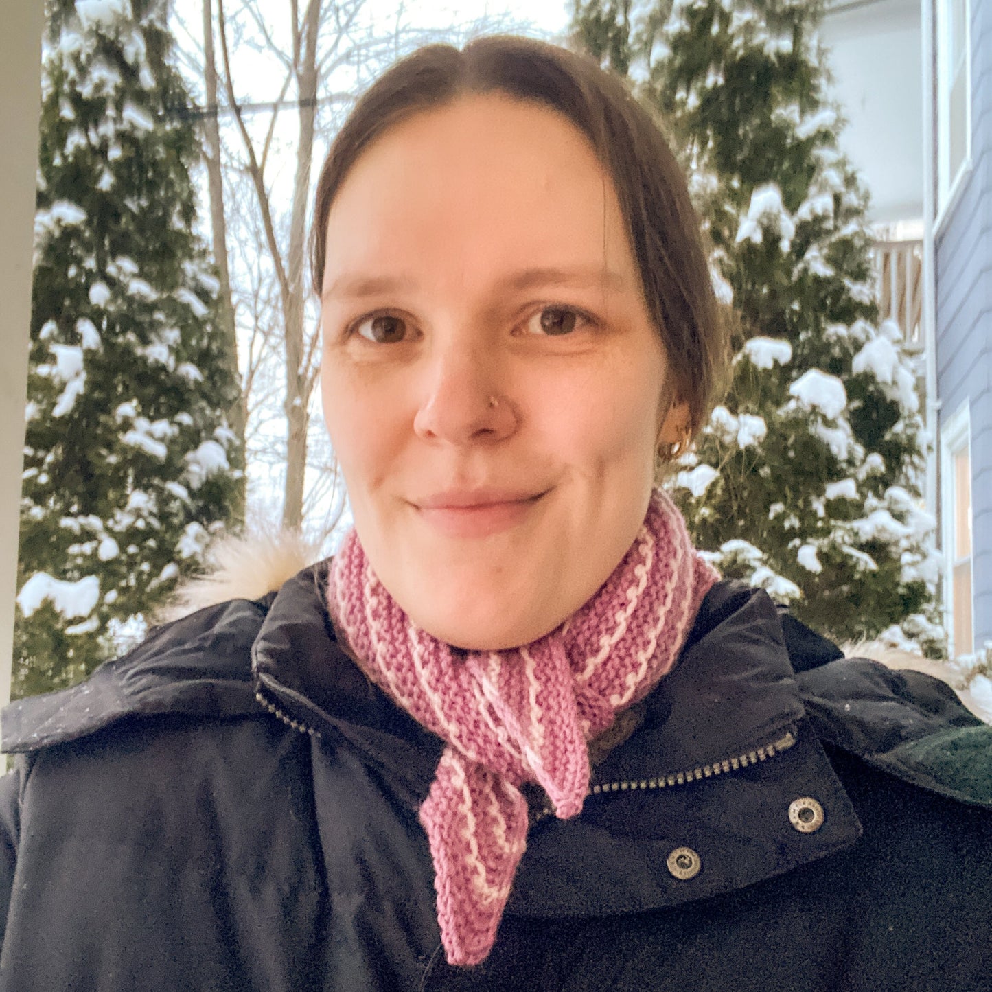 Person wearing a black coat and pink and white striped bandana outdoors with snow-covered trees in the background
