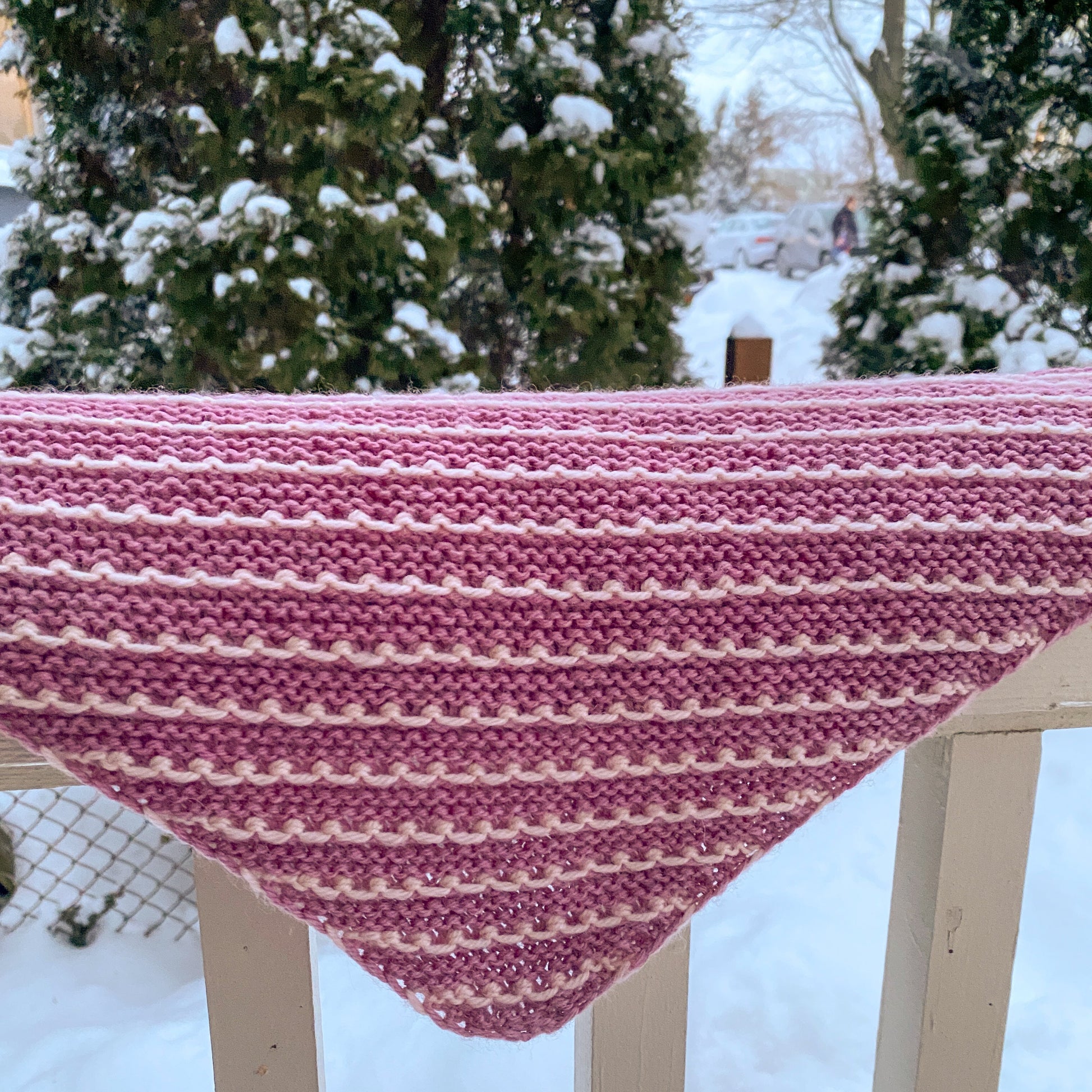 Knitted bandana scarf with pink and white pattern draped over a railing in a snowy outdoor setting.