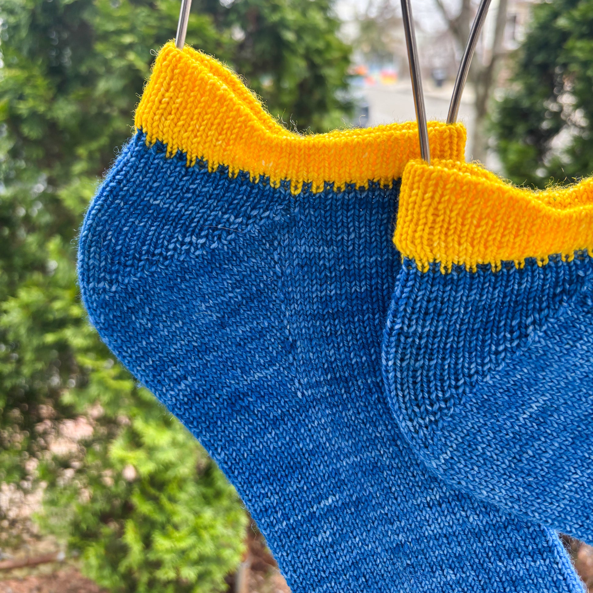 Knitted blue socks with yellow cuffs hanging on a wire against a green outdoor background