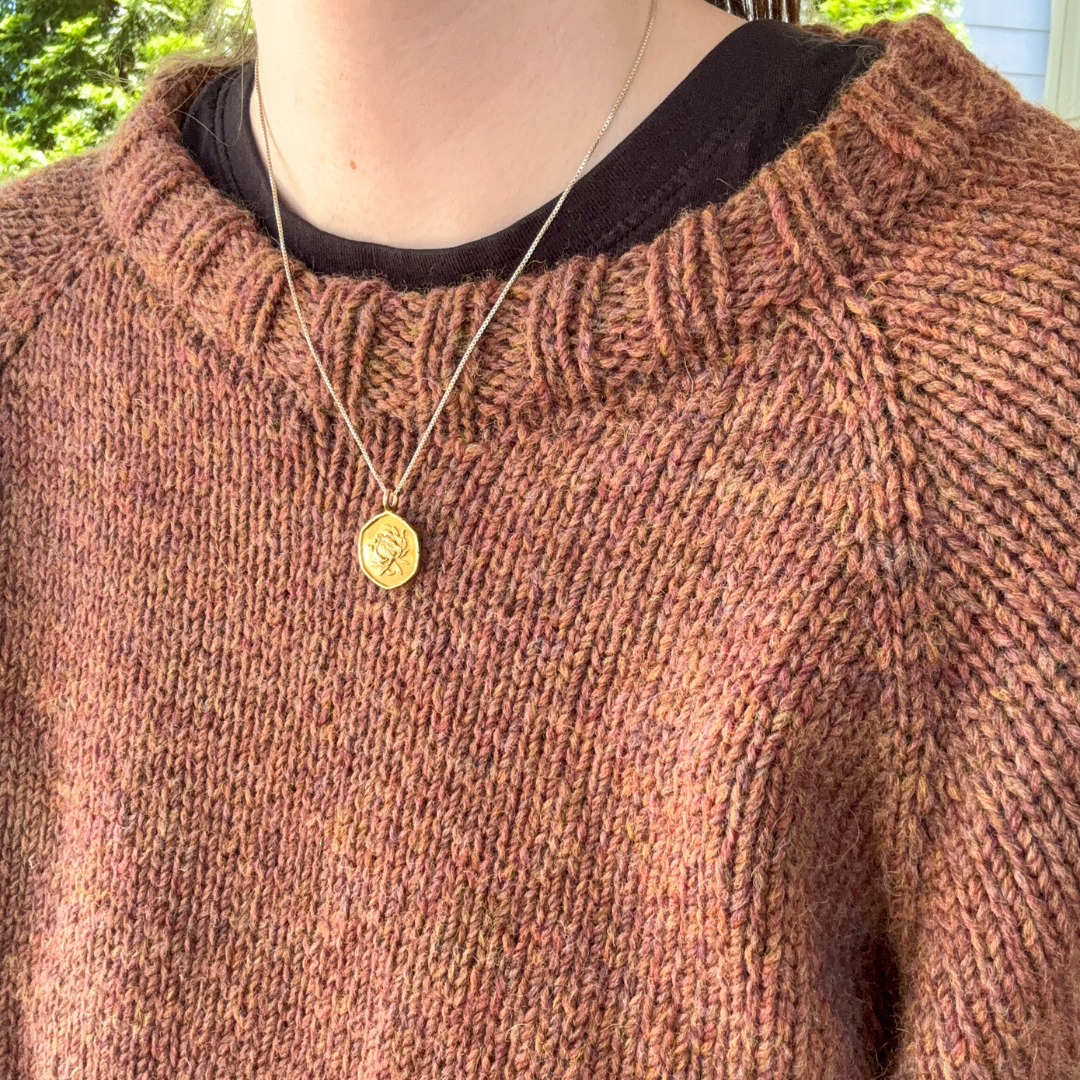 Person wearing a brown sweater with a gold necklace featuring a coin pendant.