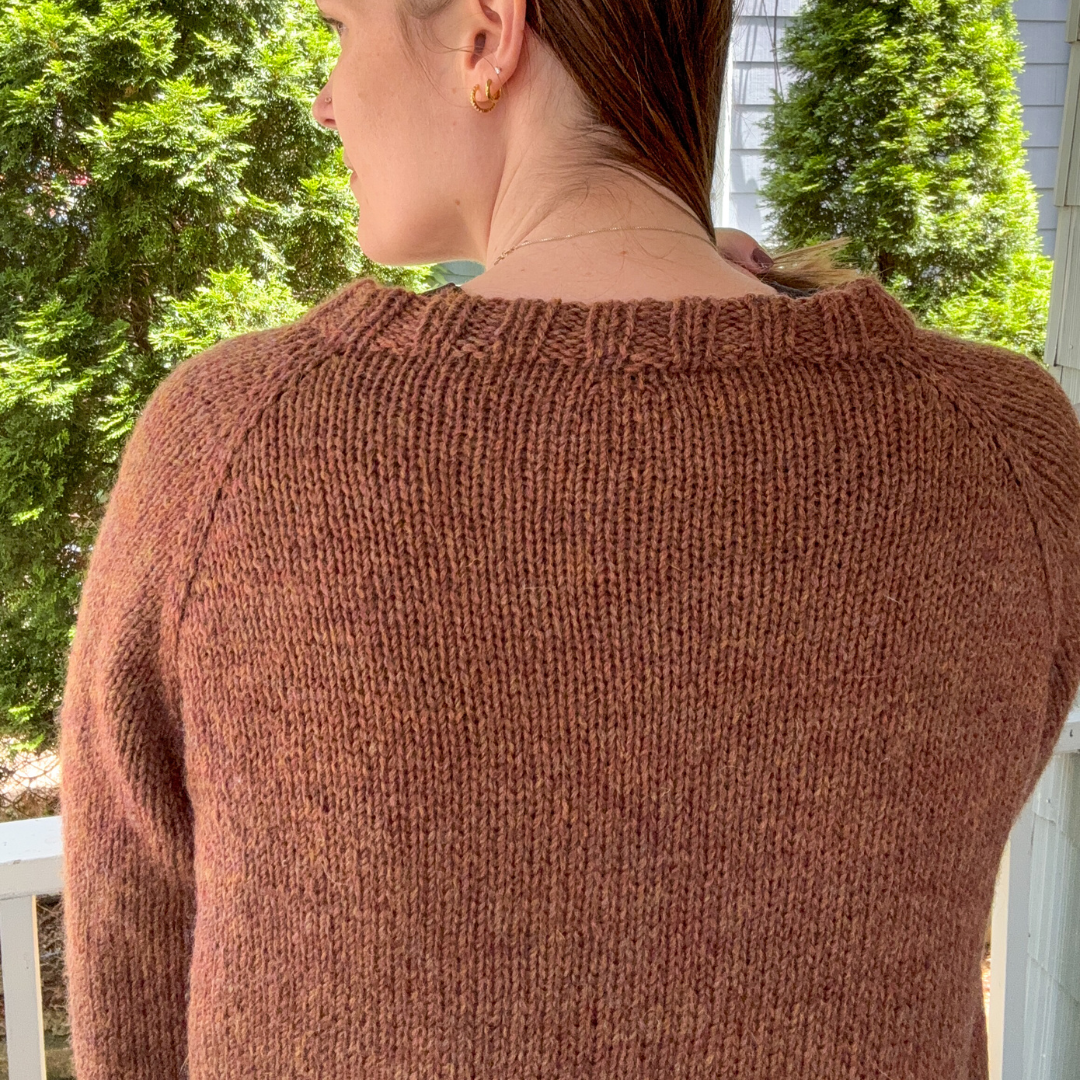 Person wearing a brown sweater with trees and a building in the background