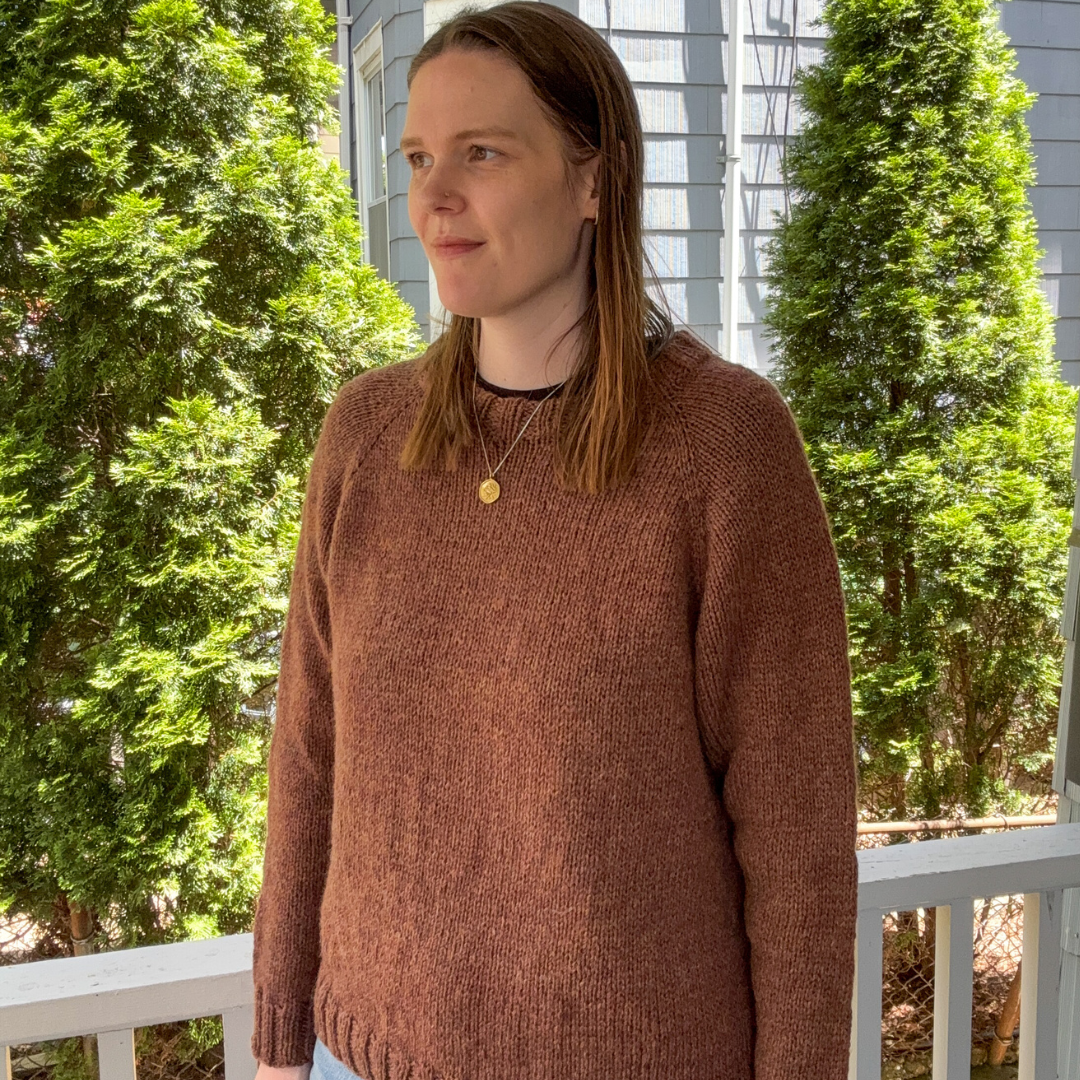 Person wearing a brown sweater standing on a porch with greenery in the background