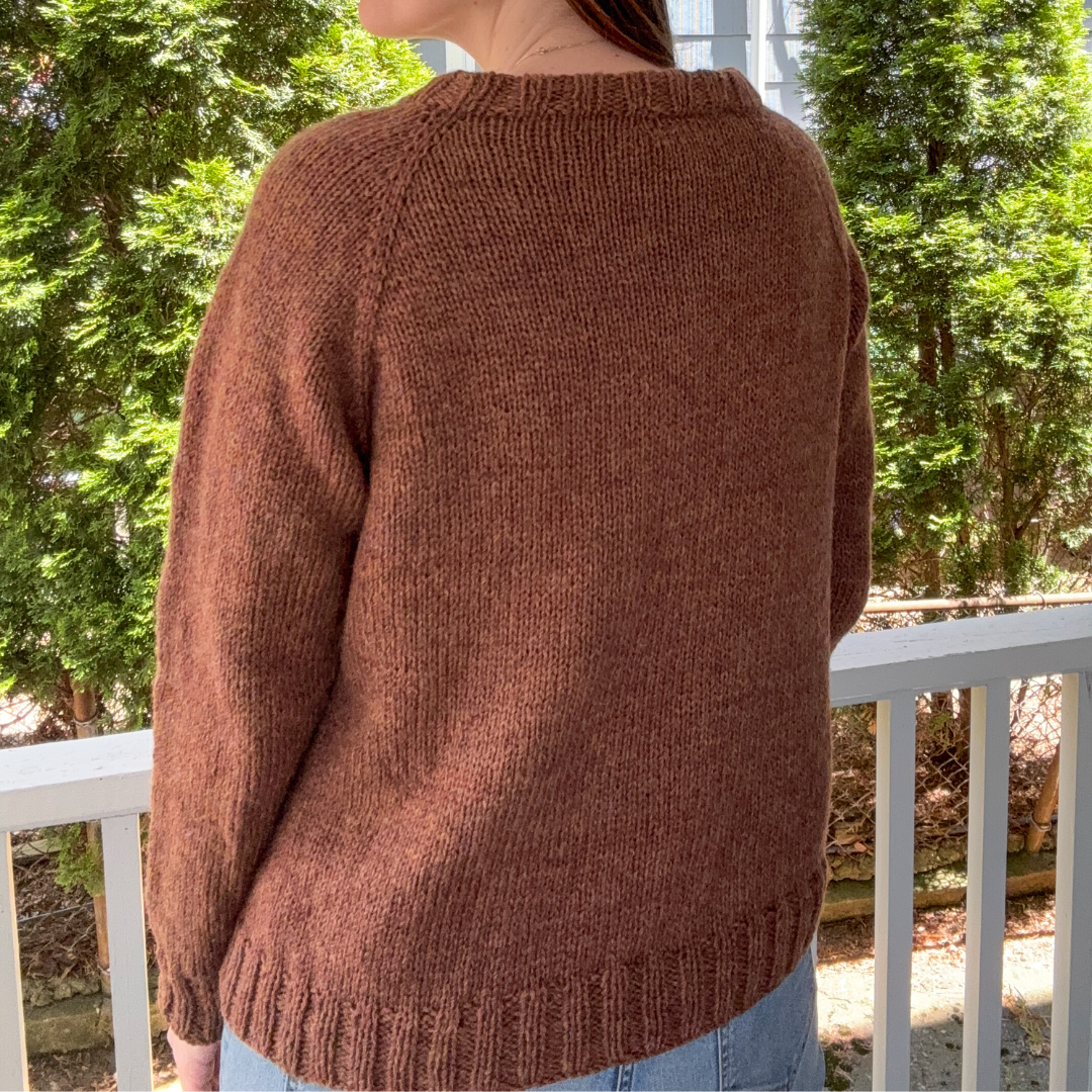 Person wearing a brown sweater standing on a balcony with trees and buildings in the background