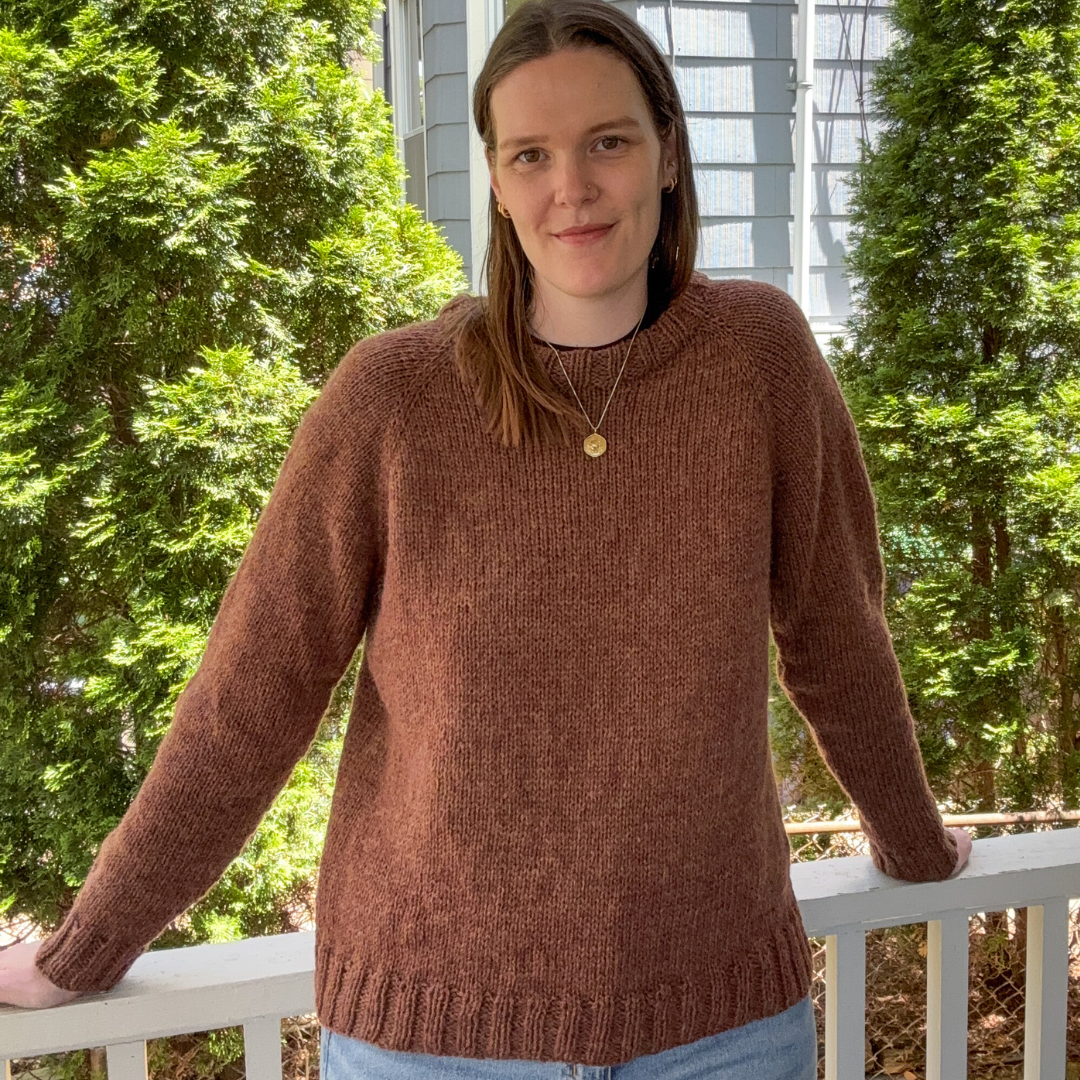 Person wearing a brown sweater and blue jeans standing on a porch with greenery in the background