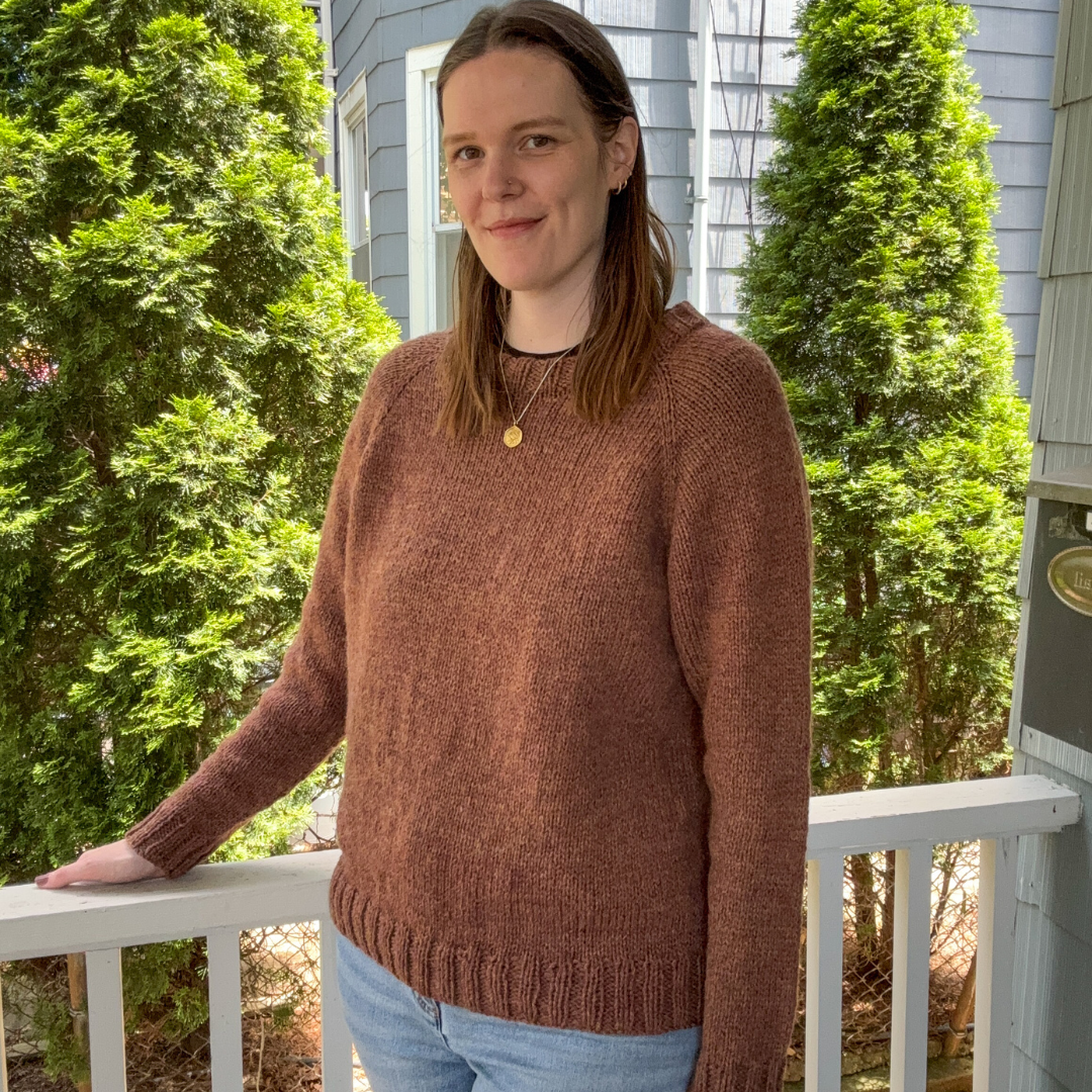 Woman wearing a brown sweater standing on a porch with greenery in the background
