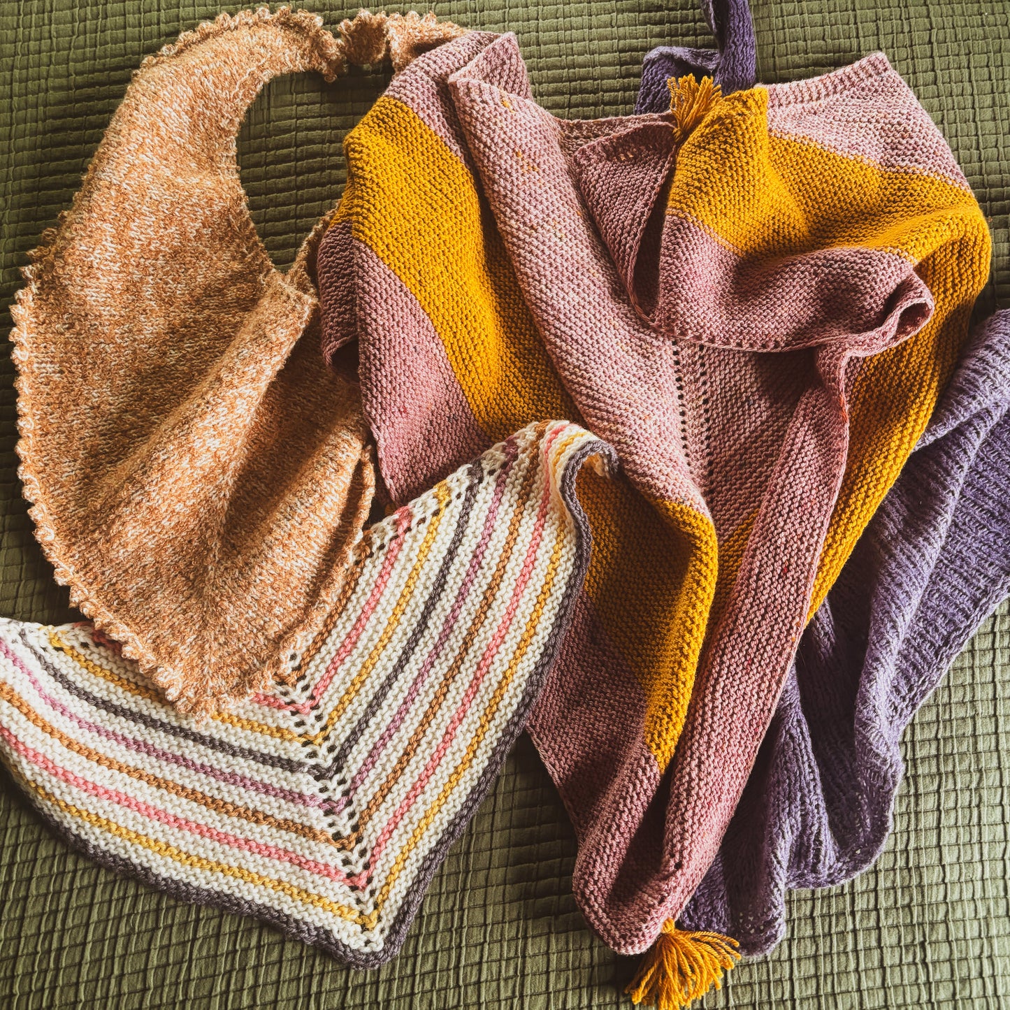 Four knitted shawls laid in a pile on a textured olive green fabric background.