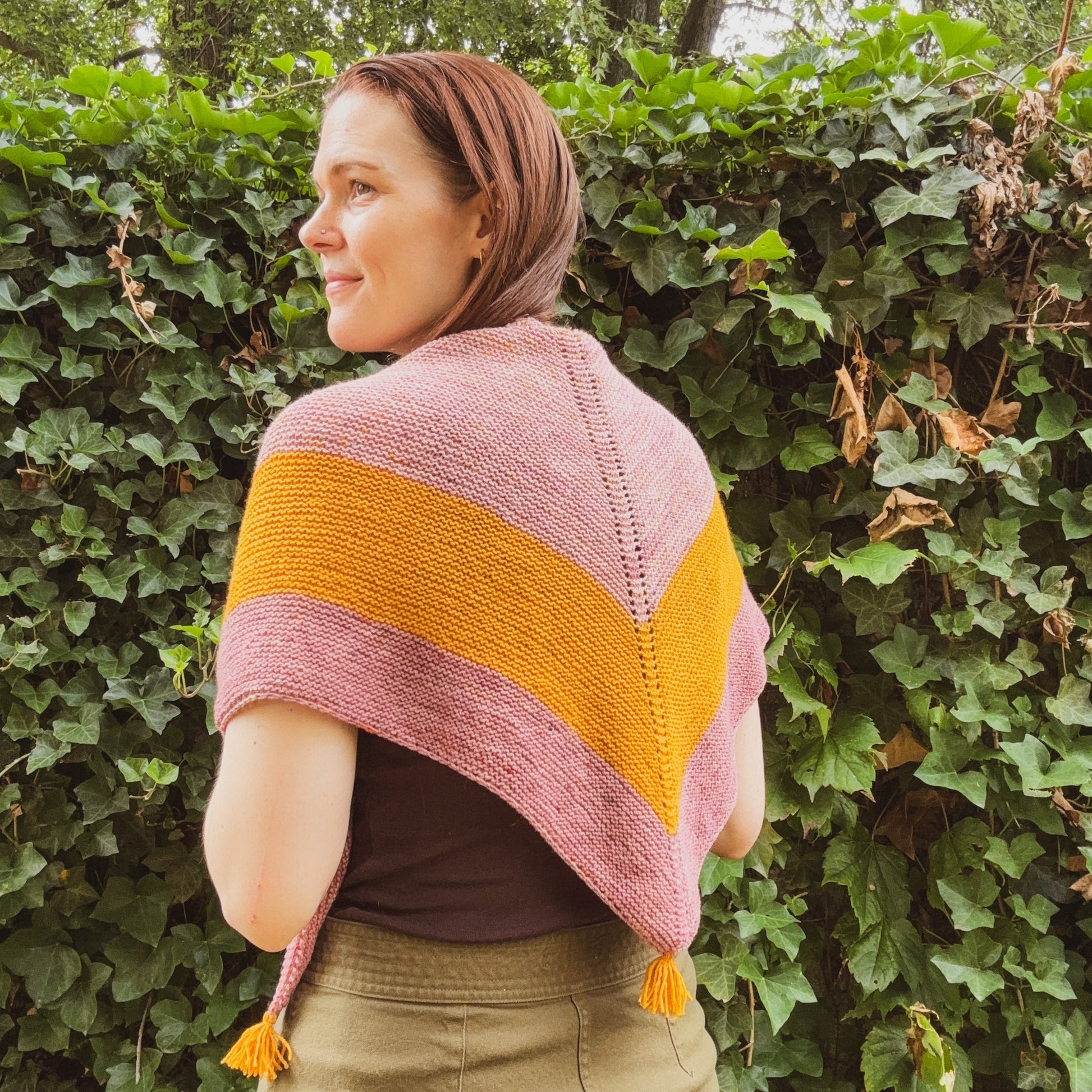 A woman wearing a hand-knit triangle shawl in dusty pink and mustard yellow, standing outdoors against a background of green ivy.
