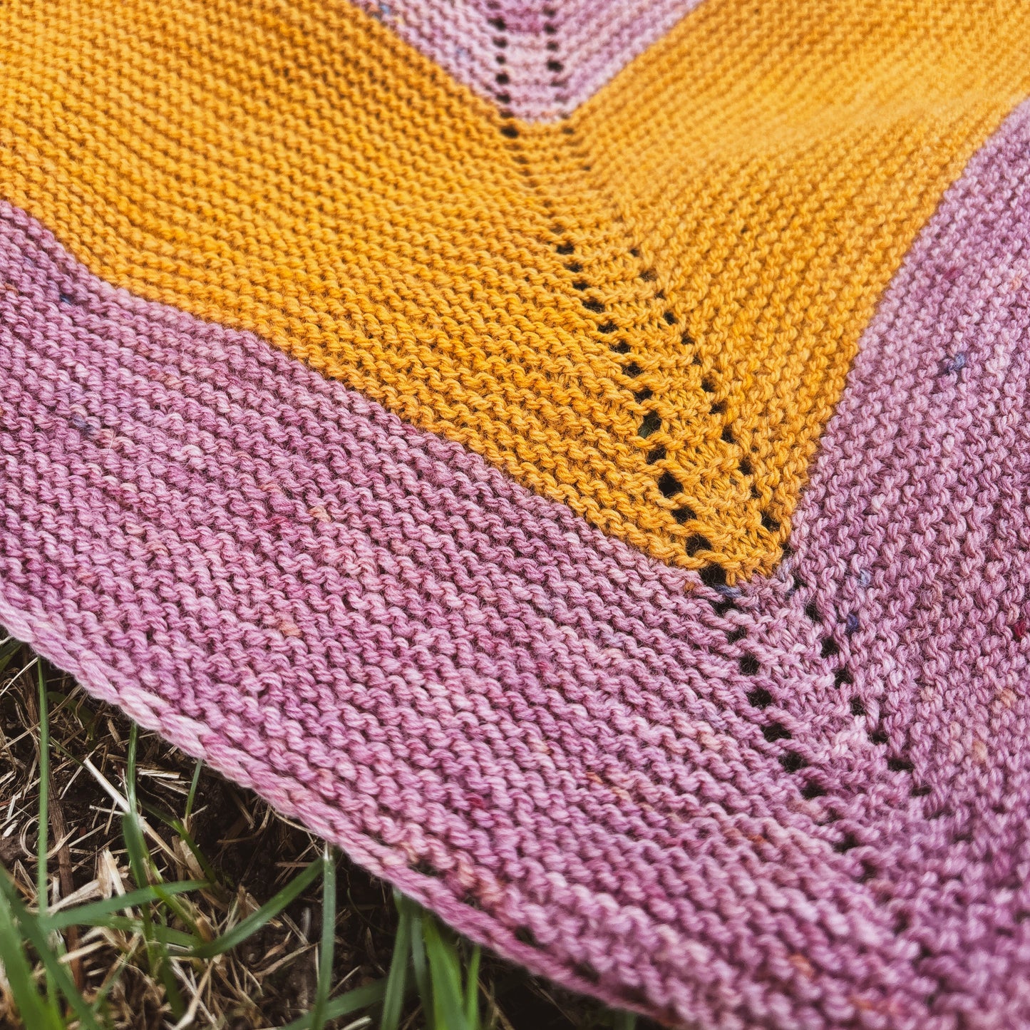 Close-up of a knitted triangle shawl with a stripe pattern in dusty pink and mustard yellow, laid outdoors on grass.
