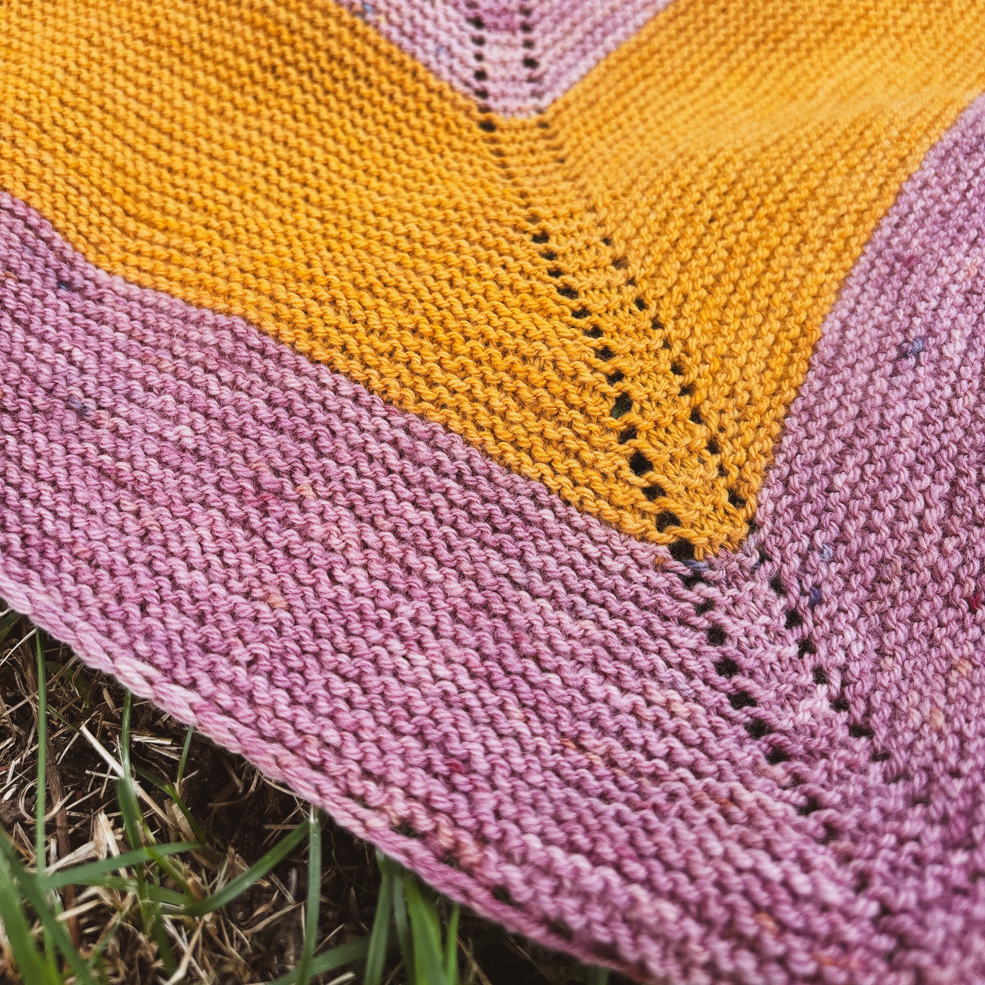 Close-up of a knitted triangle shawl with a stripe pattern in dusty pink and mustard yellow, laid outdoors on grass.
