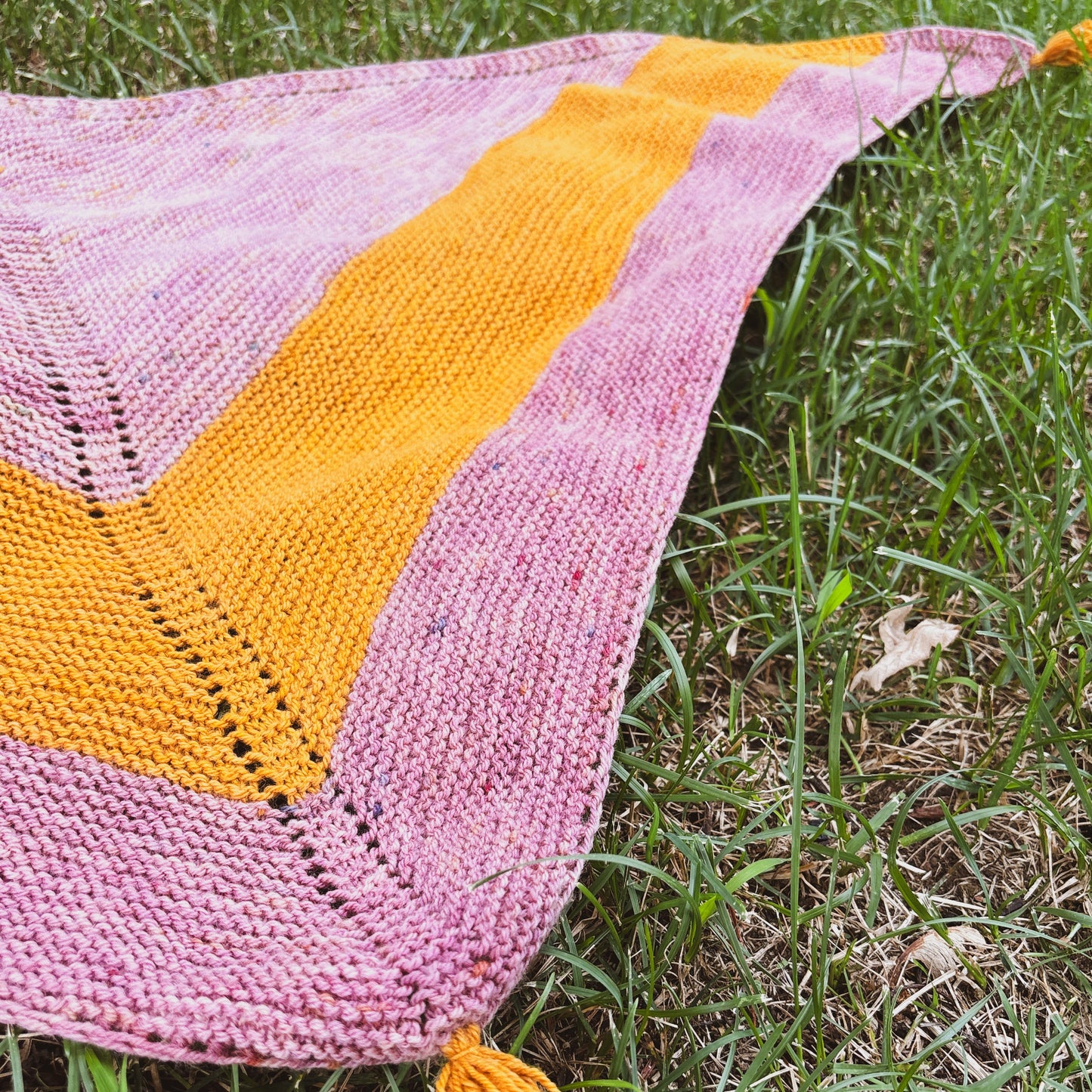 A triangular hand-knitted shawl with alternating stripes of dusty pink and mustard yellow, laid out on green grass.