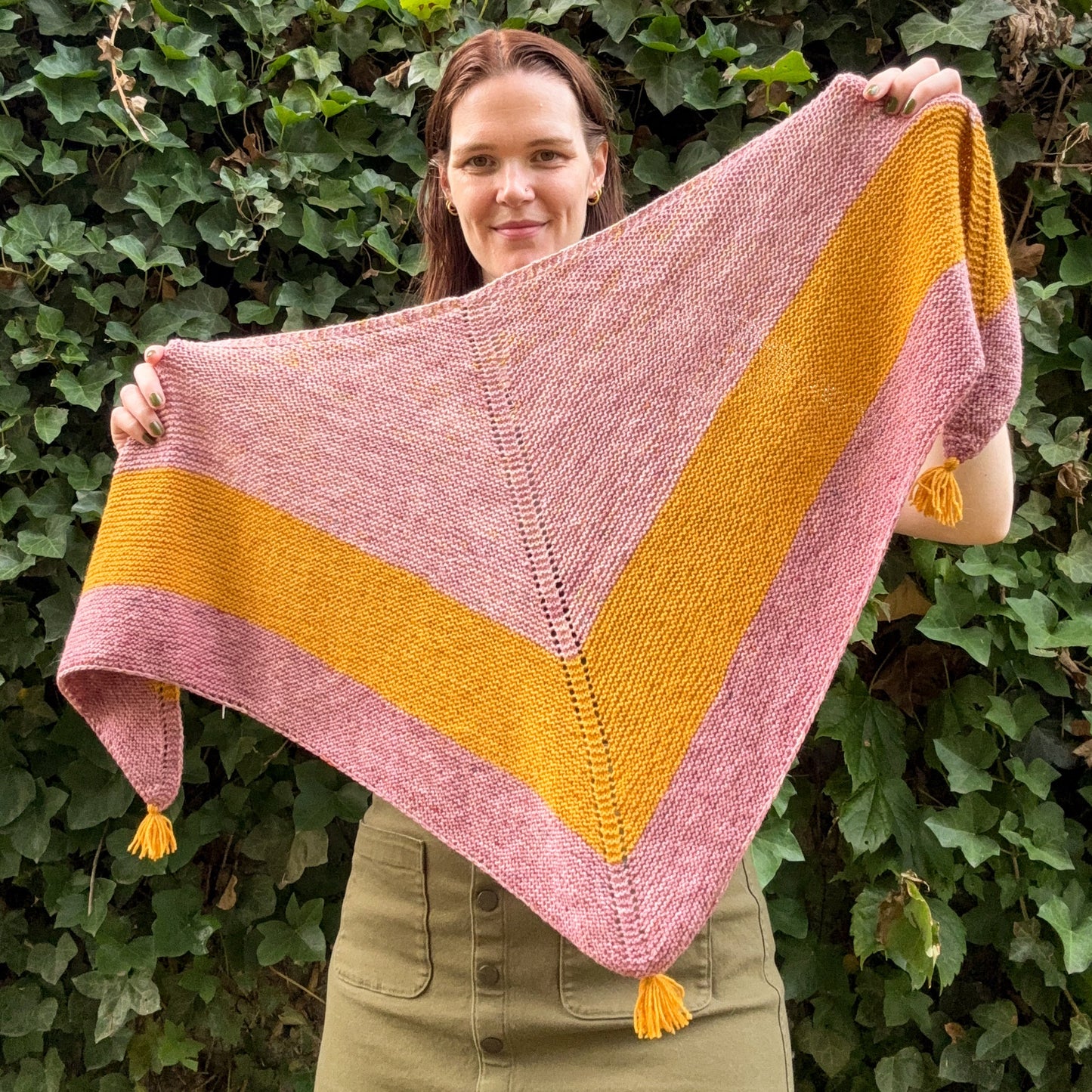 A woman outdoors in front of a green leafy hedge holds up a large triangular knitted shawl or wrap with wide pink and mustard yellow stripes and small tassels at the corners.