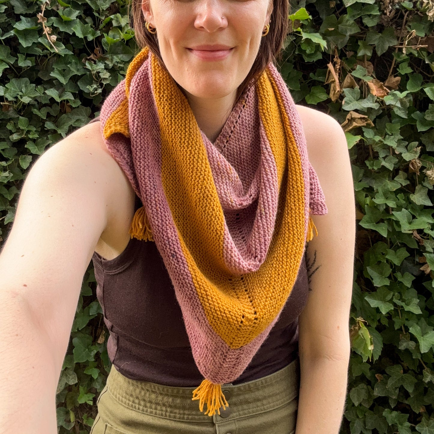 A woman wearing a knitted triangle shawl in alternating stripes of dusty rose pink and mustard yellow, standing outdoors in front of a green ivy wall.