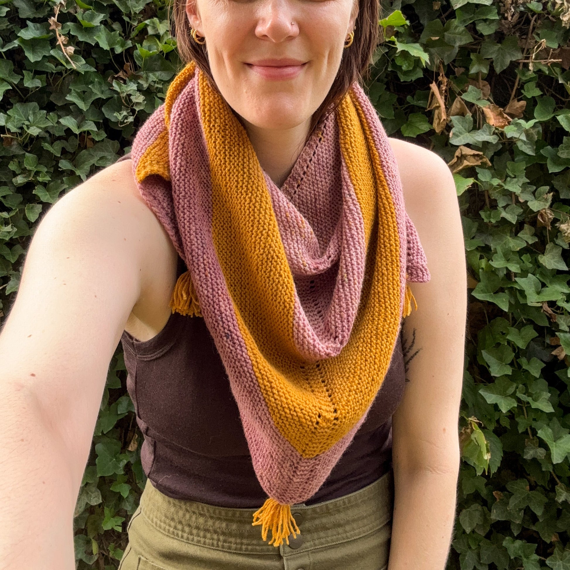 A woman wearing a knitted triangle shawl in alternating stripes of dusty rose pink and mustard yellow, standing outdoors in front of a green ivy wall.