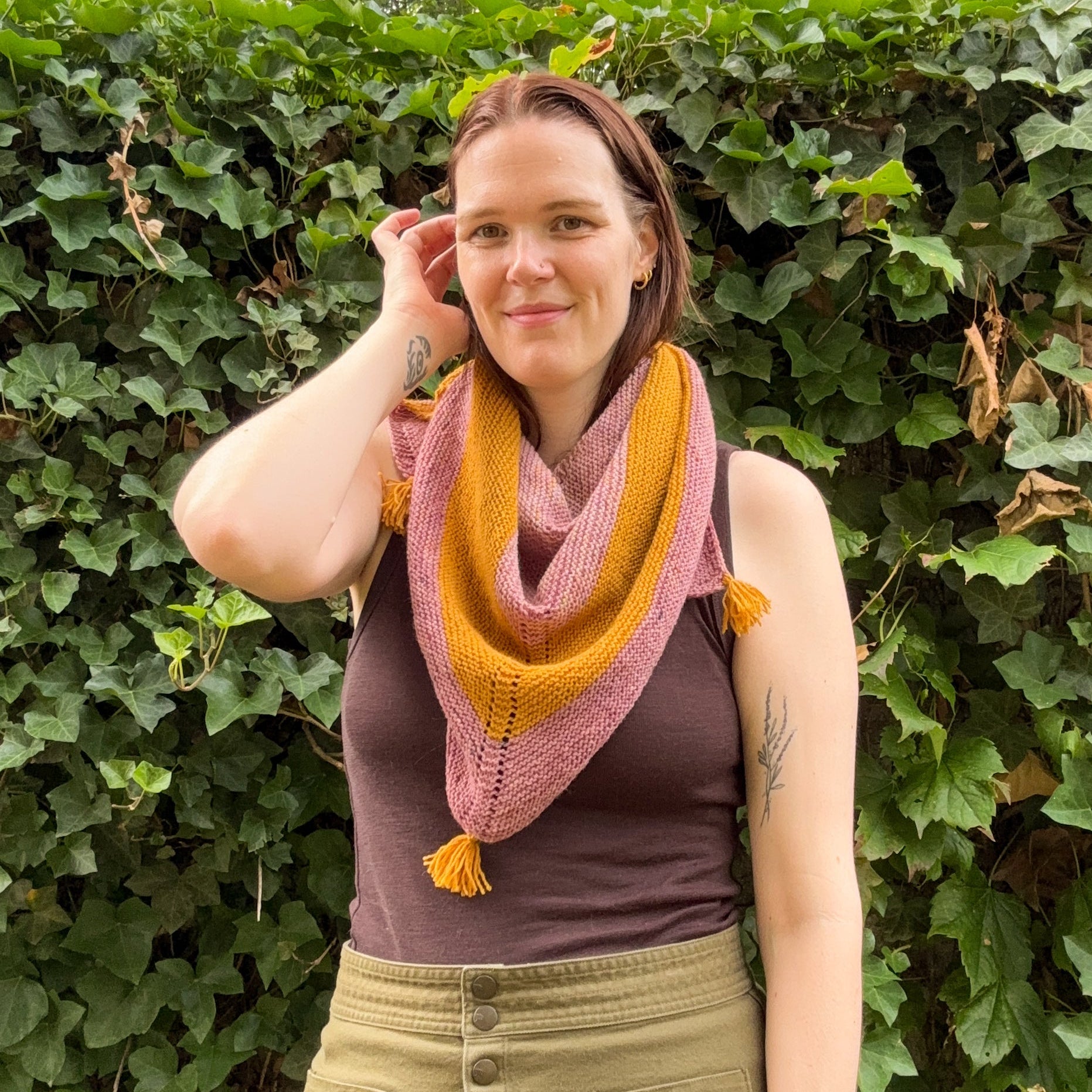 A woman wearing a pink and mustard yellow knitted shawl with tassels, standing in front of a leafy green ivy wall.