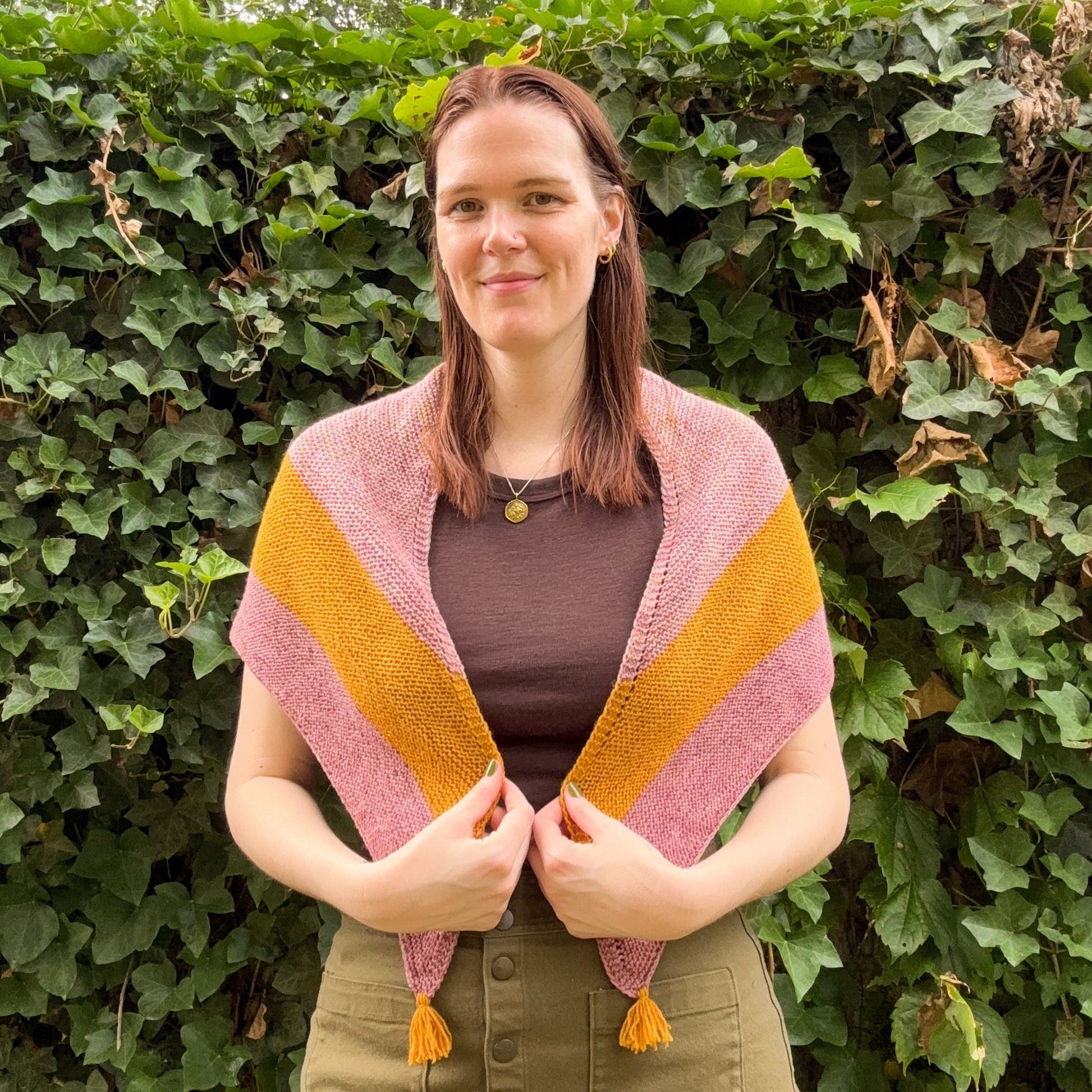 A woman wearing a pink and mustard yellow striped knit shawl with tassel ends, standing outdoors in front of a leafy green hedge.