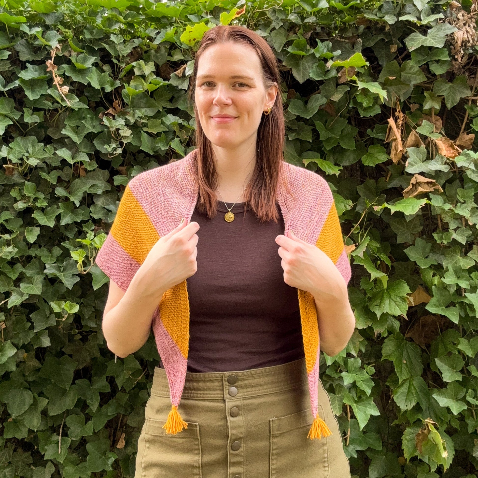 A woman wearing a pink and mustard yellow knit shawl with tassel ends, standing outdoors in front of a leafy green hedge.