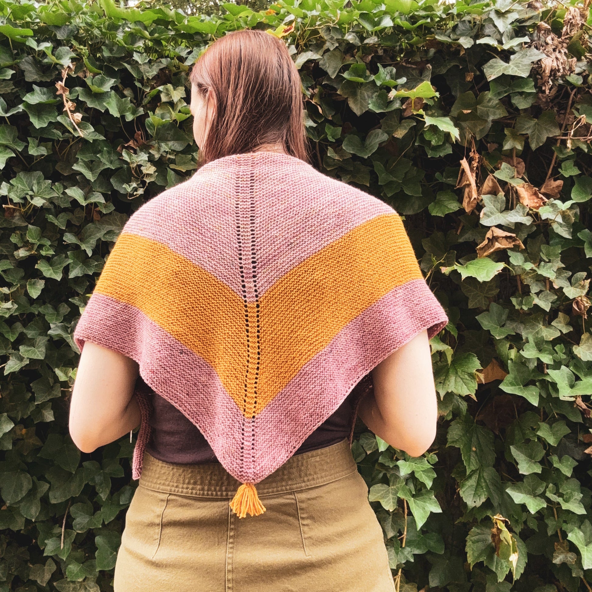A woman viewed from behind wearing a hand-knit triangular shawl with a stripe pattern in dusty pink and mustard yellow, featuring a small tassel at the bottom. She is standing outdoors in front of a dense green ivy wall.