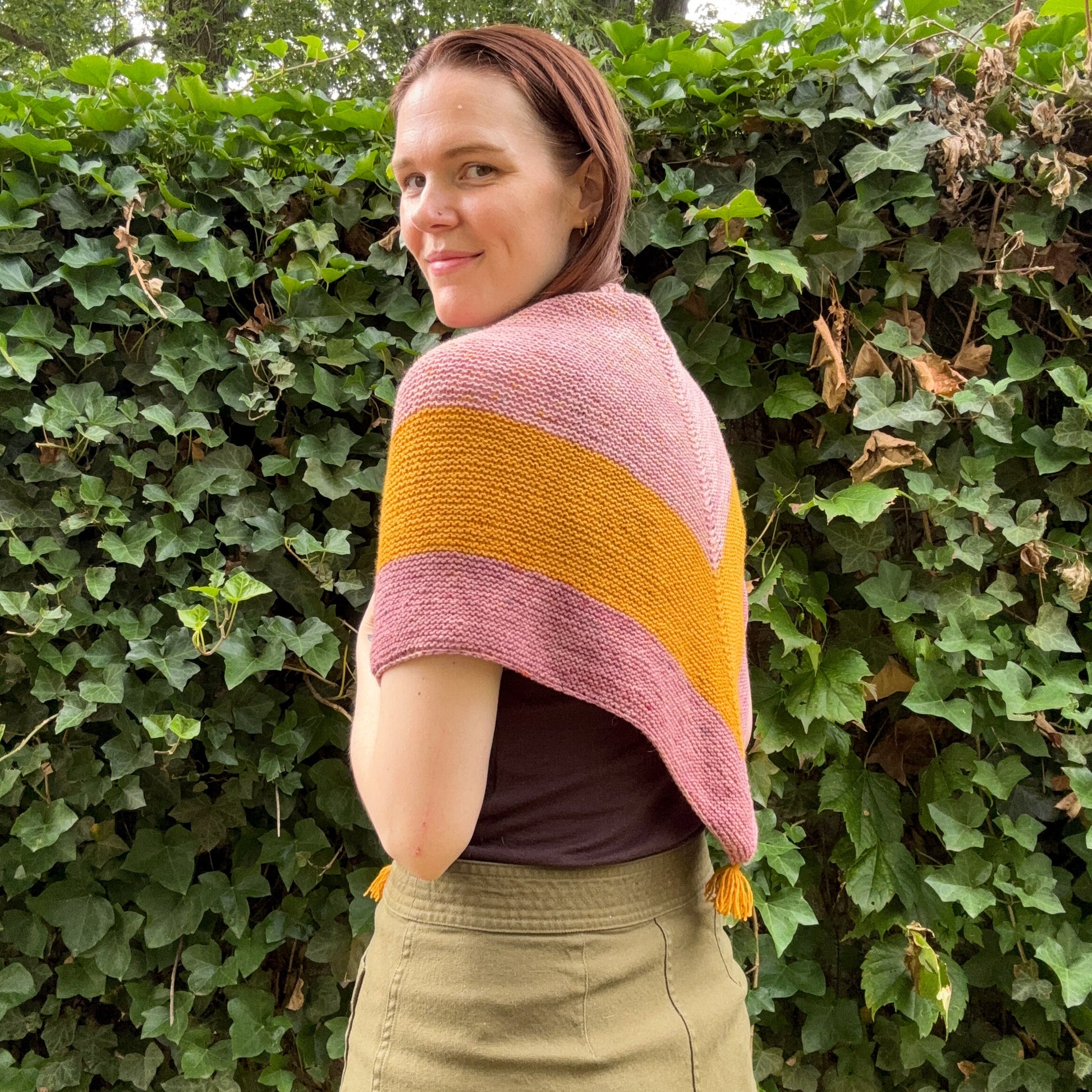A woman wearing a knit triangle shawl with stripes of dusty pink, mustard yellow, and mauve, standing outdoors against a background of green ivy.