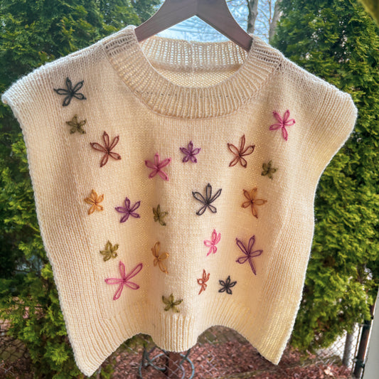 cream knitted vest with embroidered flowers on a hanger against a green outdoor background