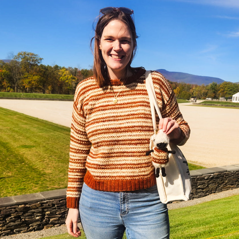 Woman in a striped knitted sweater holding a LaBaaBaa sheep charm outdoors with trees and mountains in the background