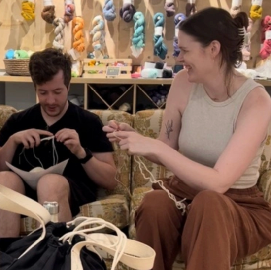 Two people sitting together in a yarn store, surrounded by colorful yarn on shelves. one is teaching the other to knit.